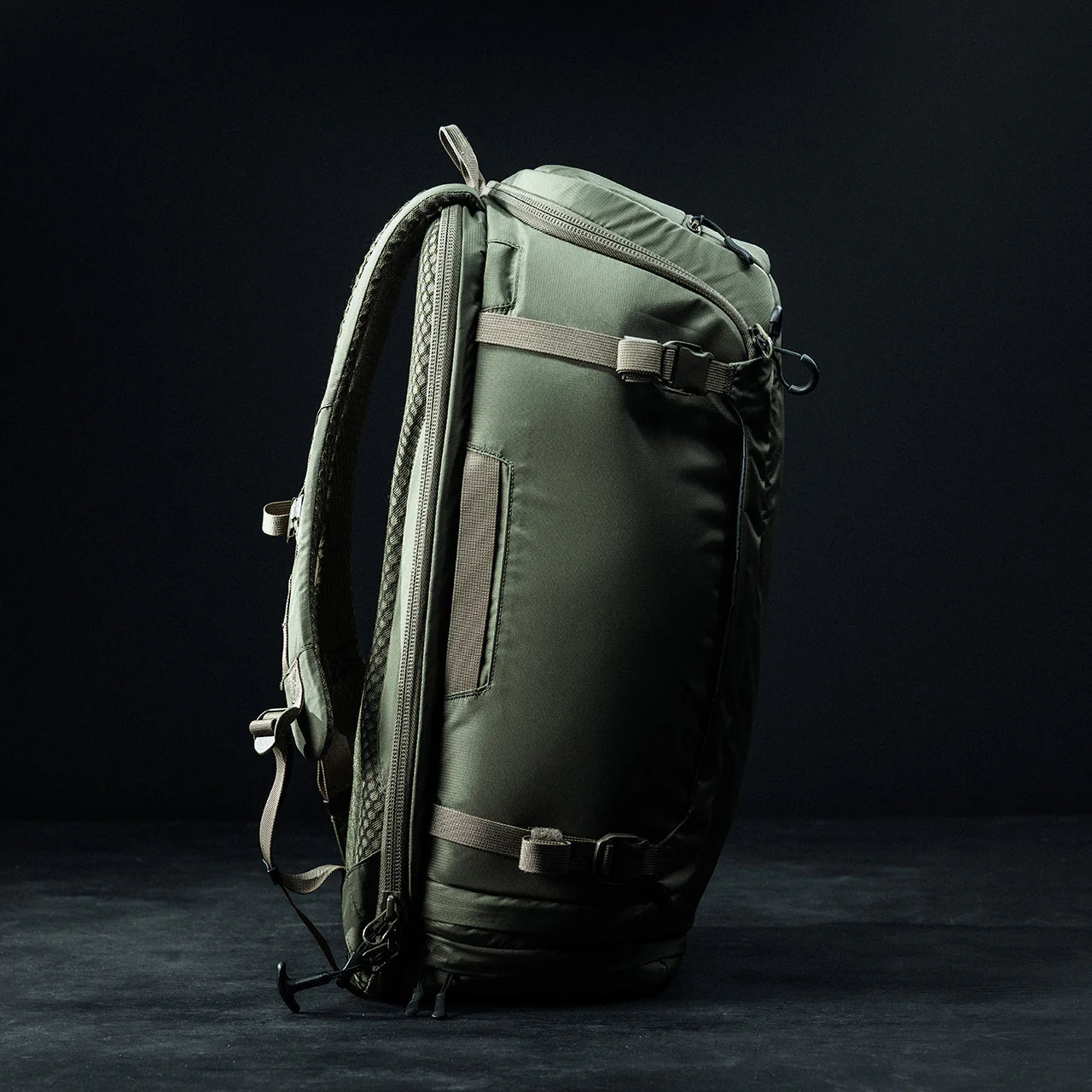Der grüne EBERLESTOCK Eberlestock Fade Adapt Rucksack steht aufrecht auf einer dunklen Fläche, von der Seite betrachtet. Er verfügt über gepolsterte Schultergurte, robuste Reißverschlüsse, mehrere verstärkte Griffe und eine integrierte ausklappbare Scheide auf schwarzem Hintergrund.