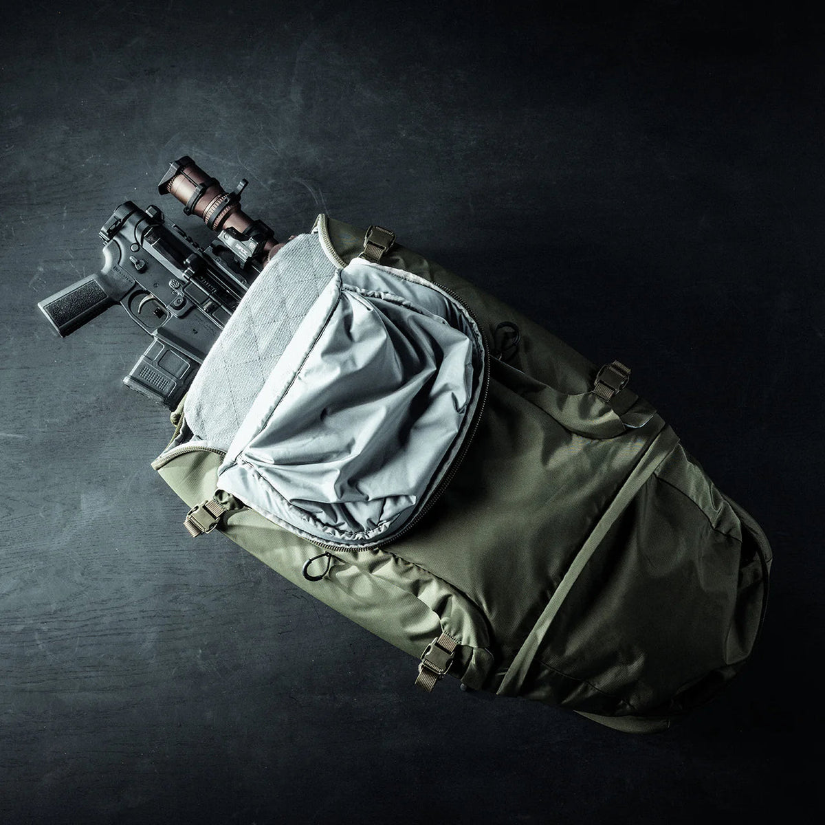 Ein EBERLESTOCK Eberlestock Fade Adapt liegt geöffnet auf einer dunklen Fläche und enthüllt ein schwarzes Gewehr mit einem braunen Zielfernrohr, das teilweise in das Hauptfach gepackt ist.