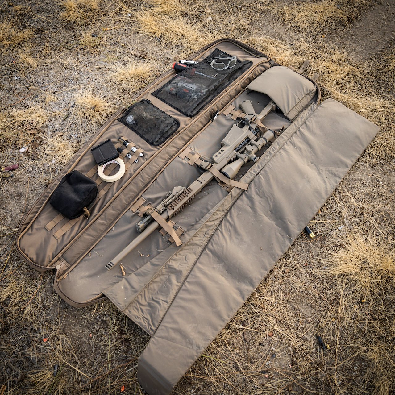 Ein EBERLESTOCK Sniper Sled Drag Bag 52" liegt offen auf trockenem Gras und zeigt ein zerlegtes Gewehr, eine schwarze Tasche, Klebeband, Gehörschutz, Schutzbrille und kleines Zubehör, das ordentlich in Netztaschen angeordnet ist.