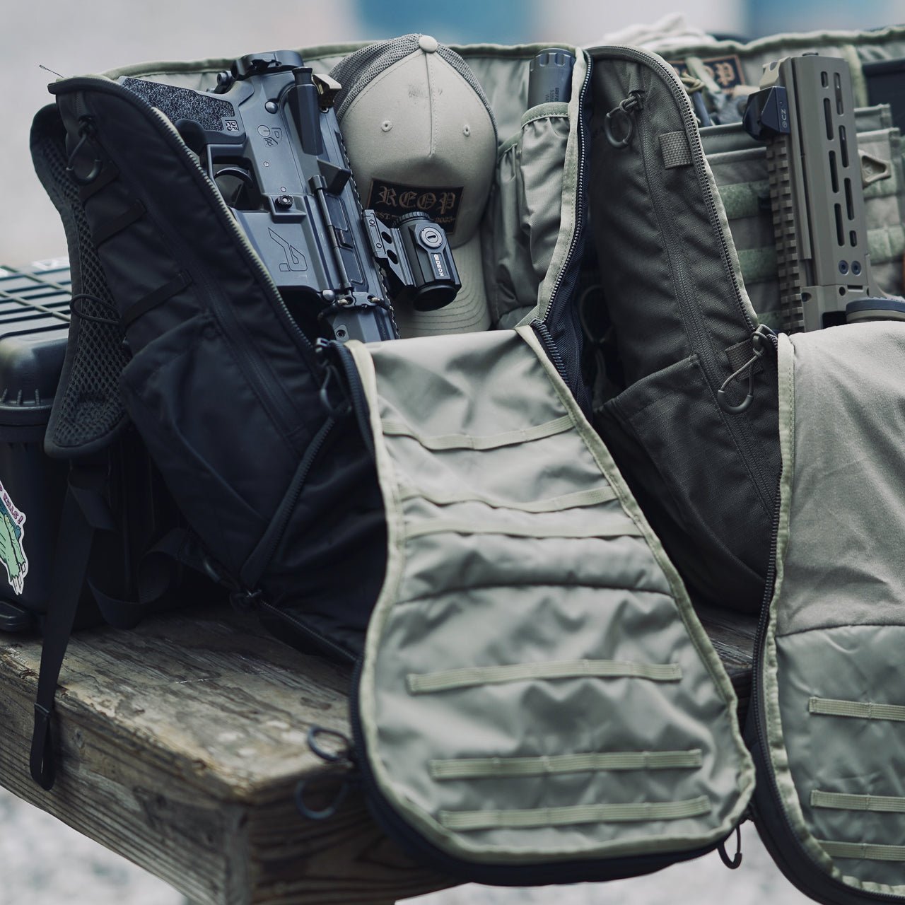 Ein EBERLESTOCK Little Trick Pack liegt mit geöffnetem Reißverschluss auf einem Holztisch und zeigt Gewehr und Ausrüstung im Inneren. Eine andere in gedämpftem Grün und Schwarz, mit einem versteckten Waffenfach und einer Tasche, deutet auf eine Schieß- oder Trainingsausrüstung hin.
