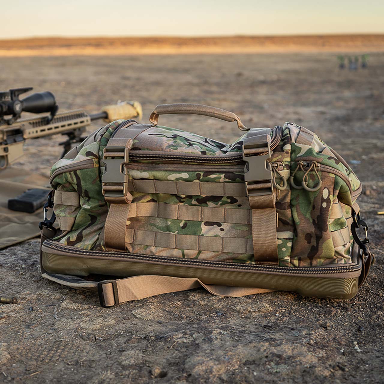 Ein EBERLESTOCK Bang Bag in Tarnfarben mit braunen Riemen liegt auf sandigem Boden neben einem Gewehr und einer braunen Matte in der offenen Landschaft eines Schießstandes unter klarem Himmel.