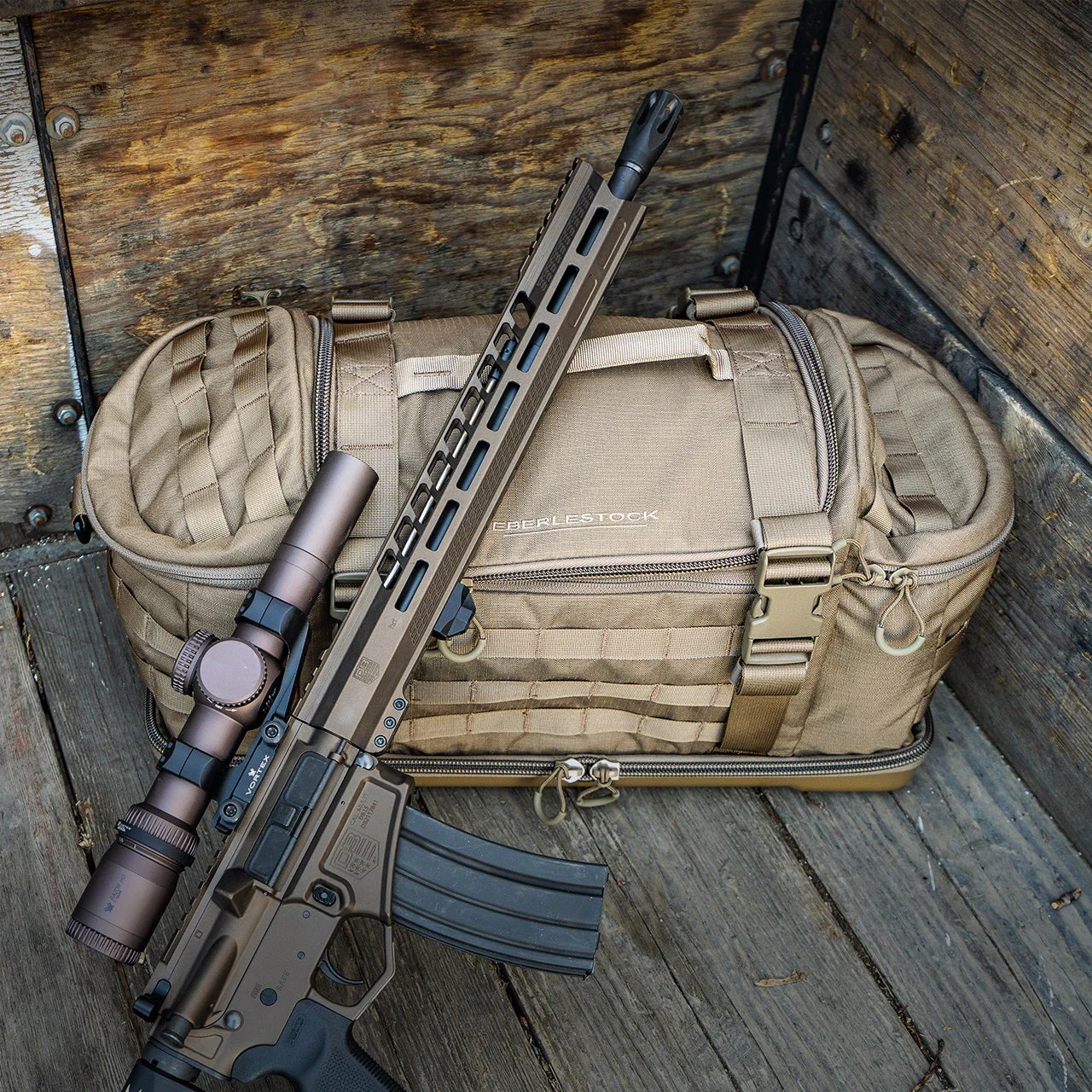 Eine EBERLESTOCK Bang Bag Schießstandtasche liegt auf einer Holzfläche, an der schräg ein hellbraunes und schwarzes Gewehr mit Zielfernrohr lehnt - mit MOLLE-Gurtband und Reißverschluss, ideal für den Schießstand oder den nächsten Schießtag.