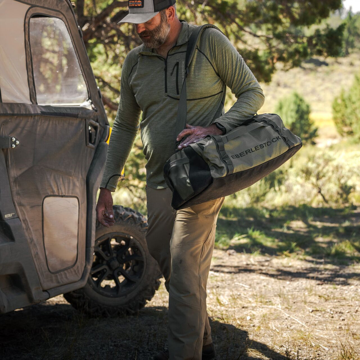 Ein Mann in Outdoor-Ausrüstung trägt einen EBERLESTOCK Hyllus Duffel 65 und steht neben einem Geländewagen auf einer sonnenbeschienenen, von Bäumen umgebenen Wiese.