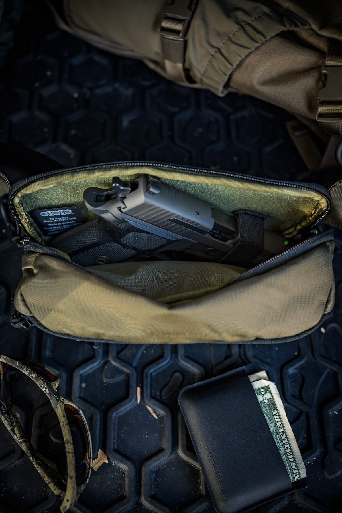 Eine EBERLESTOCK Bando Bag Standard liegt offen auf einer schwarzen, strukturierten Oberfläche, in der eine Handfeuerwaffe sichtbar ist. In der Nähe befinden sich eine Sonnenbrille und eine schwarze Brieftasche, in der Bargeld teilweise sichtbar ist.
