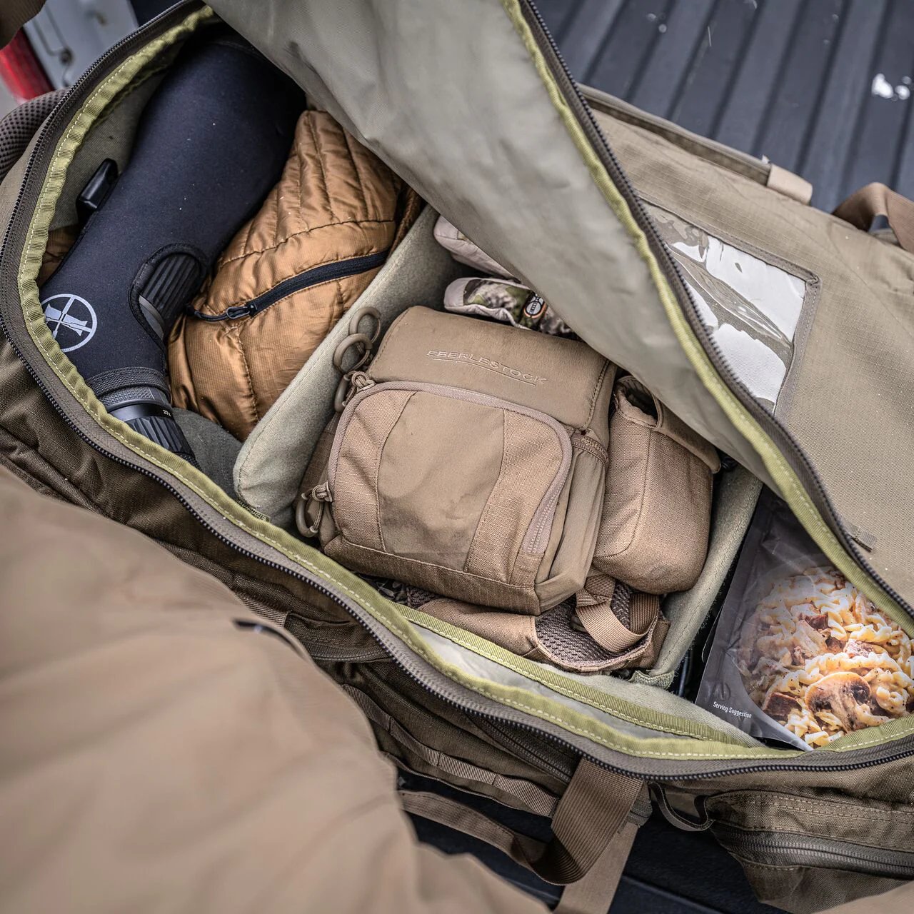 Ein großer khakifarbener EBERLESTOCK Atlas Duffel offenbart eine gut organisierte Outdoor-Ausrüstung, darunter ein Zielfernrohr, eine braune Jacke, Taschen und ein Essensbehälter in der robusten Reisetasche.