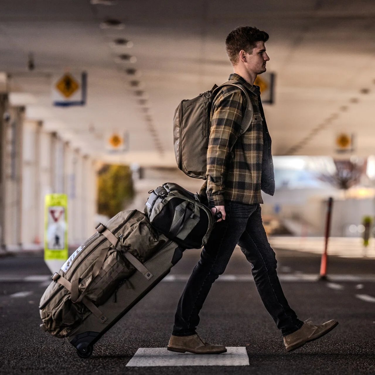Ein Mann in einem karierten Hemd, Jeans und Stiefeln geht mit einem Rucksack ins Freie und zieht einen EBERLESTOCK Atlas Duffel unter einen überdachten Bereich, wahrscheinlich an einem Flughafen oder einem Bahnhof.