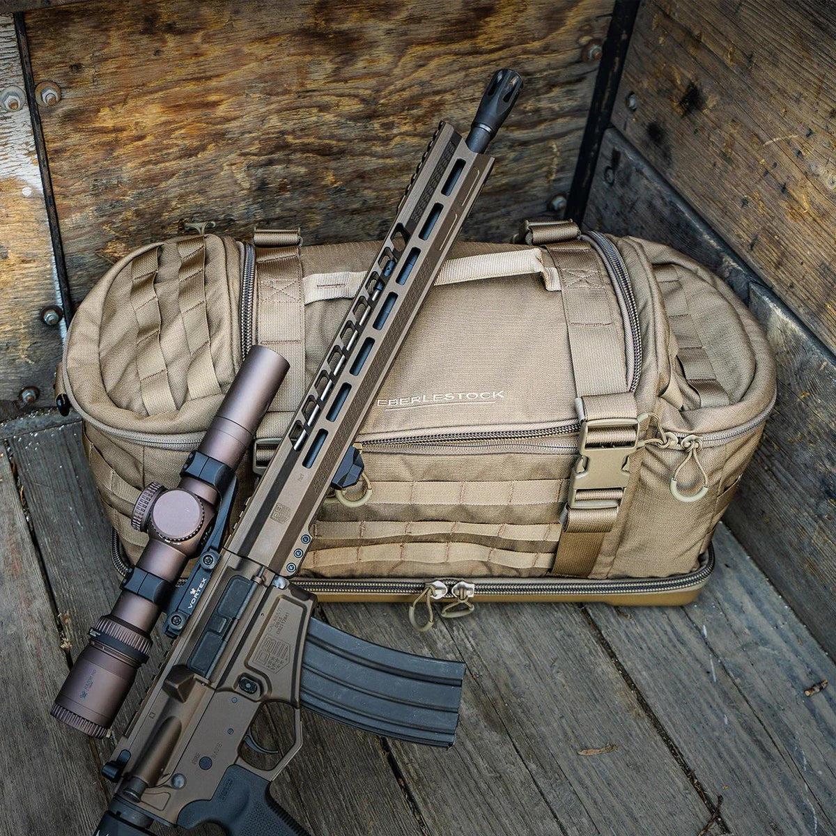 Ein braunes taktisches Gewehr mit Zielfernrohr lehnt an einer EBERLESTOCK Bang Bag Schießsporttasche auf einer hölzernen Unterlage in einer Kiste, bereit für den Einsatz am Schießstand.