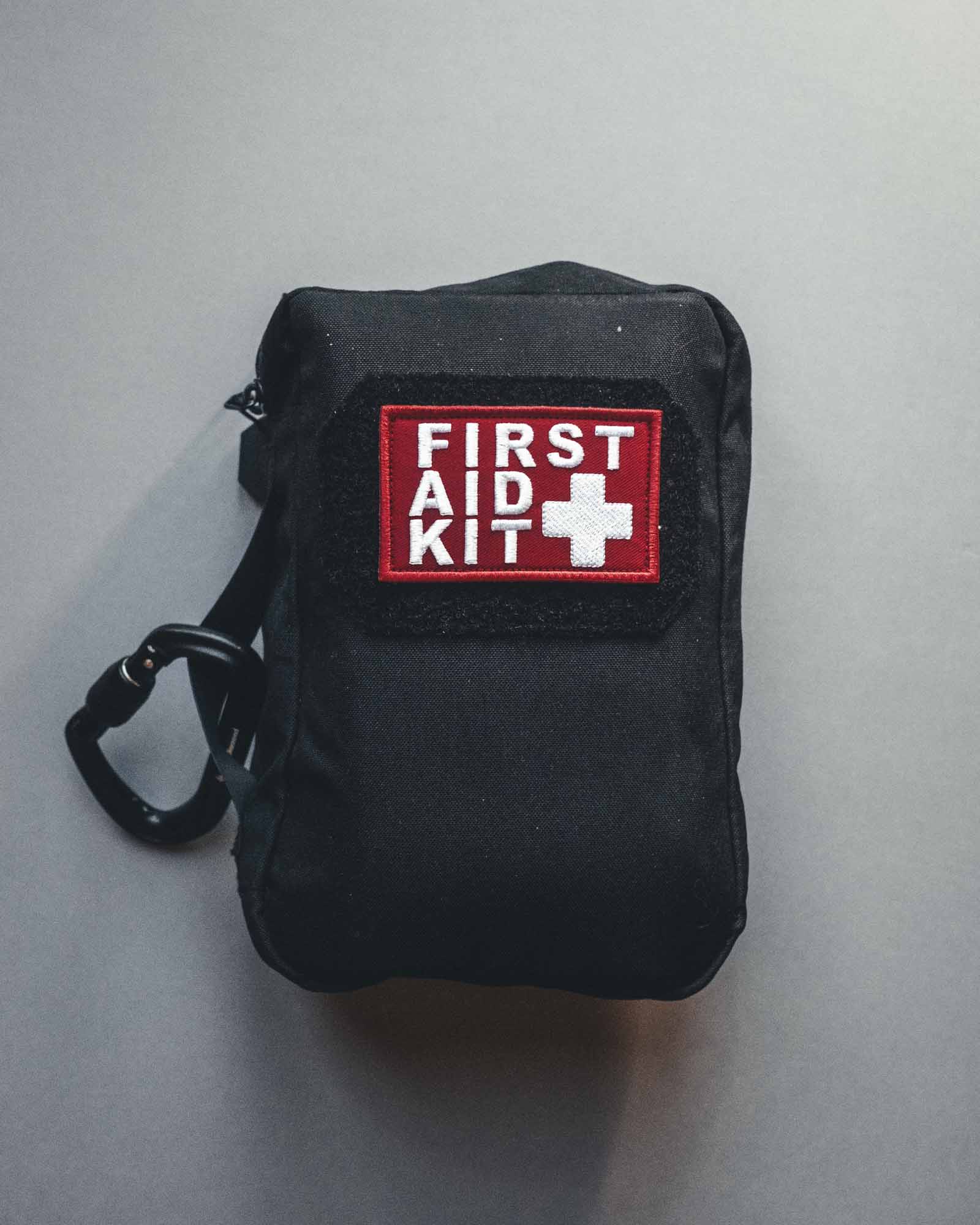 Die STOIRM Large Pouch V.2, ein schwarzes Erste-Hilfe-Set von STOIRM mit rot-weißem "FIRST AID KIT"-Aufnäher und weißem Kreuz, ist Molle-Panel-kompatibel und steht auf einem schlichten grauen Hintergrund.