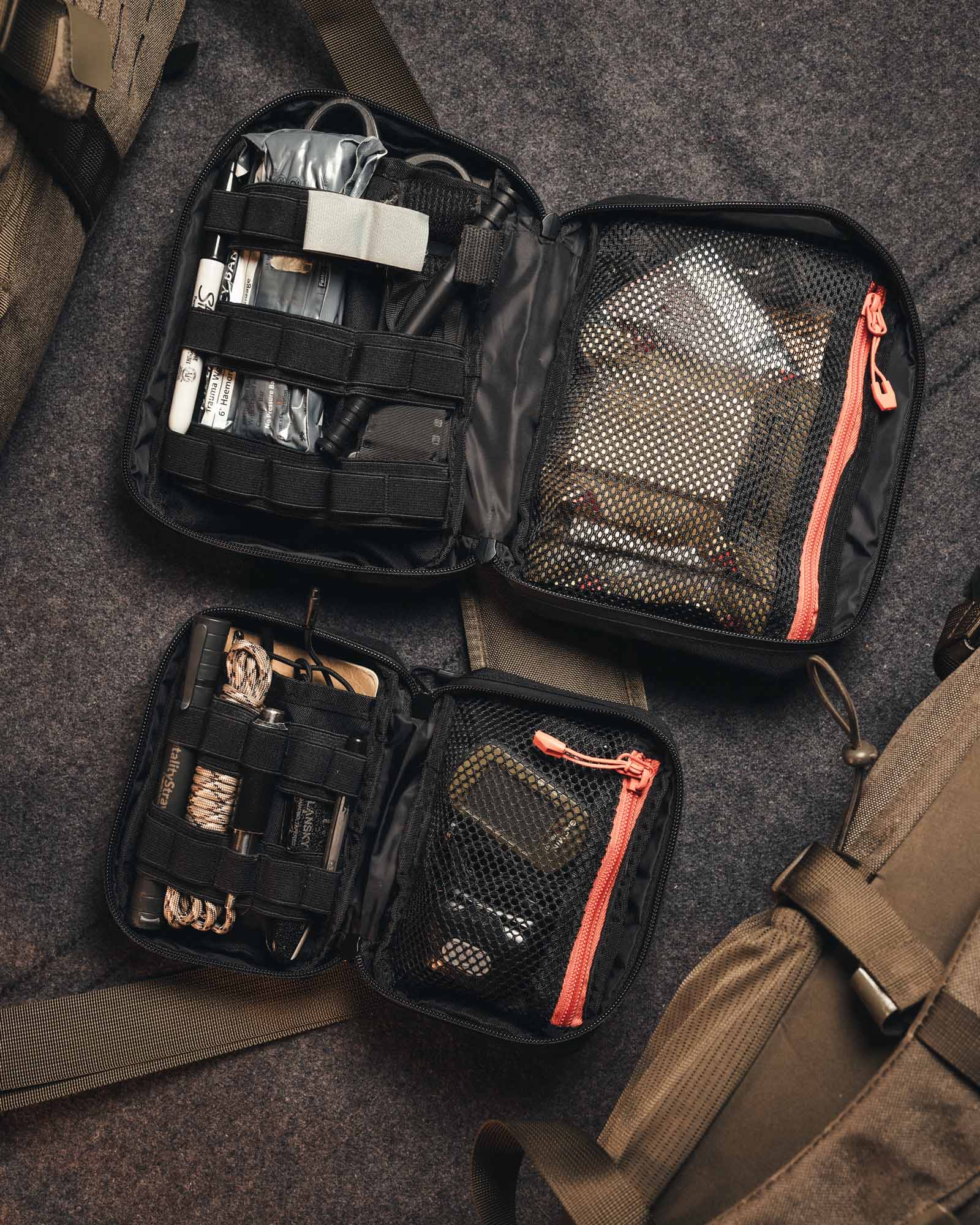 Zwei offene schwarze Utensilientaschen, darunter eine STOIRM Small Pouch V.2 mit Netztaschen und elastischen Riemen, nehmen kleine Werkzeuge und Vorräte auf. Sie werden auf einer grauen Stoffoberfläche neben braunen Taschen oder Rucksäcken ausgestellt.