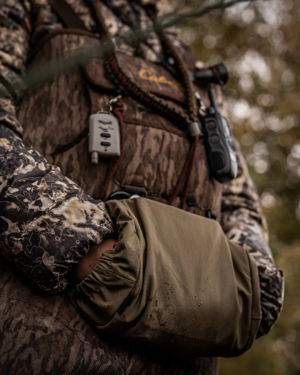 Ein Jäger in voller Tarnkleidung hält seine Hände in einem EBERLESTOCK Recon Handwarmer. In der Weste befindet sich Zubehör wie ein Funkgerät und ein Binogeschirr, während im Hintergrund unscharfes natürliches Laub zu sehen ist.