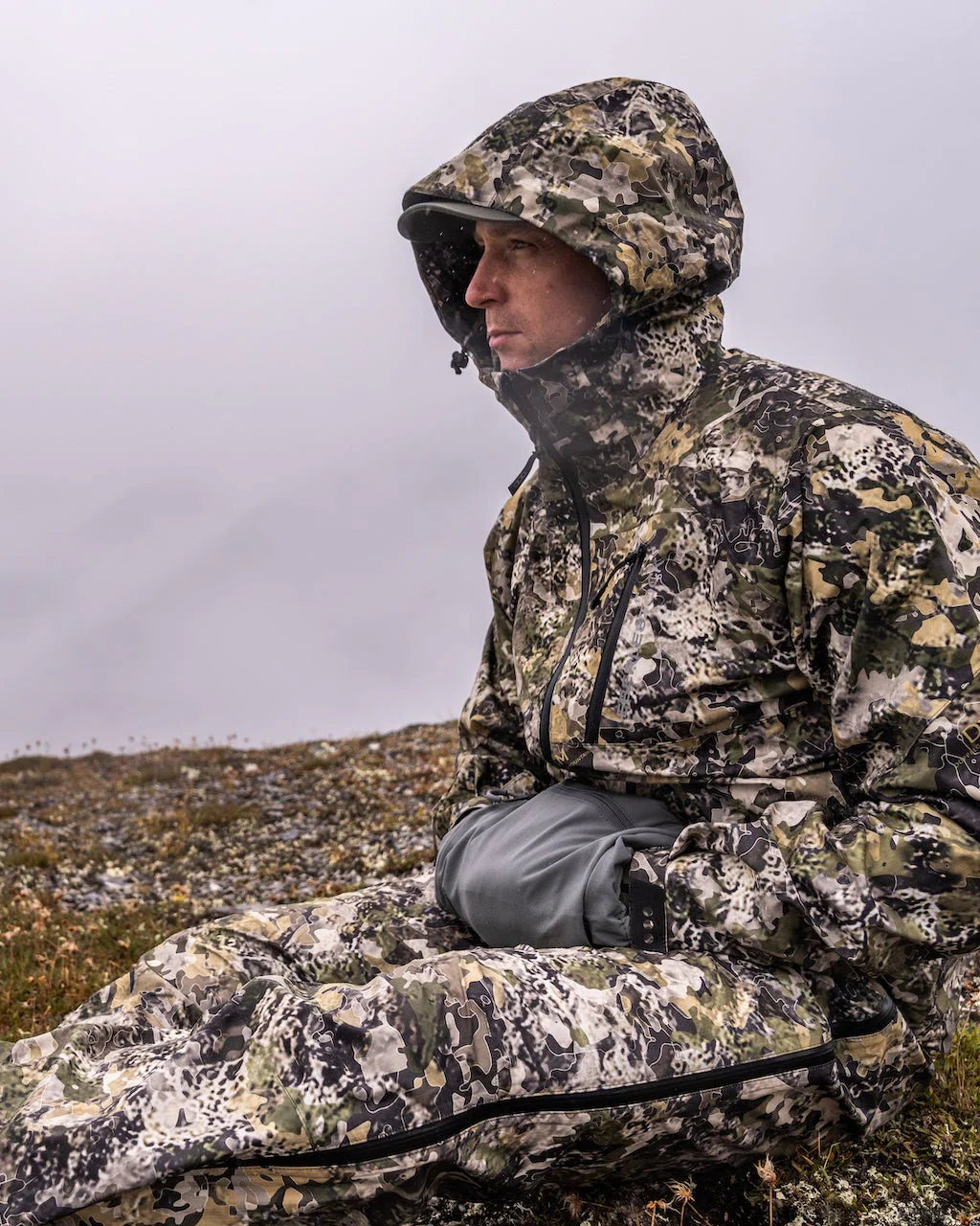 Eine Person, die eine Tarnkapuzenjacke und -hose mit einem EBERLESTOCK Recon Handwarmer trägt, sitzt an einem nebligen oder bewölkten Tag im Freien auf felsigem Boden und blickt in die Ferne.