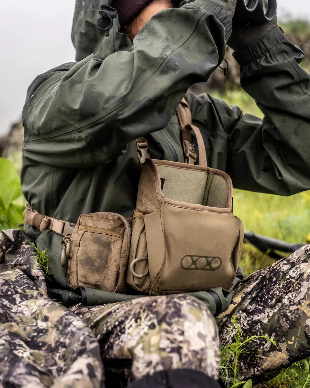Eine Person in Tarnhose und grüner Jacke sitzt im Freien, trägt die EBERLESTOCK Recon Utility Pouch in tan mit MOLLE-kompatiblem Design und hält ein Fernglas, während sie möglicherweise Wildtiere beobachtet oder jagt.