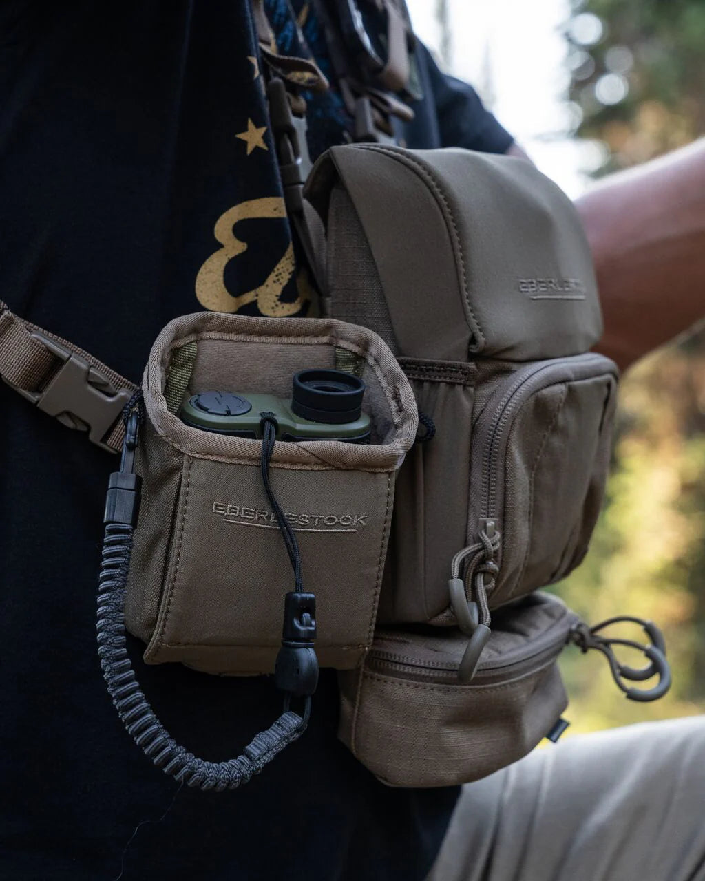 Die EBERLESTOCK Recon Rangefinder Pouch in hellbraun, die für Ihr Entfernungsmessgerät und Ihre Leine konzipiert wurde, lässt sich sicher an Ihrem taktischen Brustgeschirr befestigen - hier im Freien über einem schwarzen Hemd.
