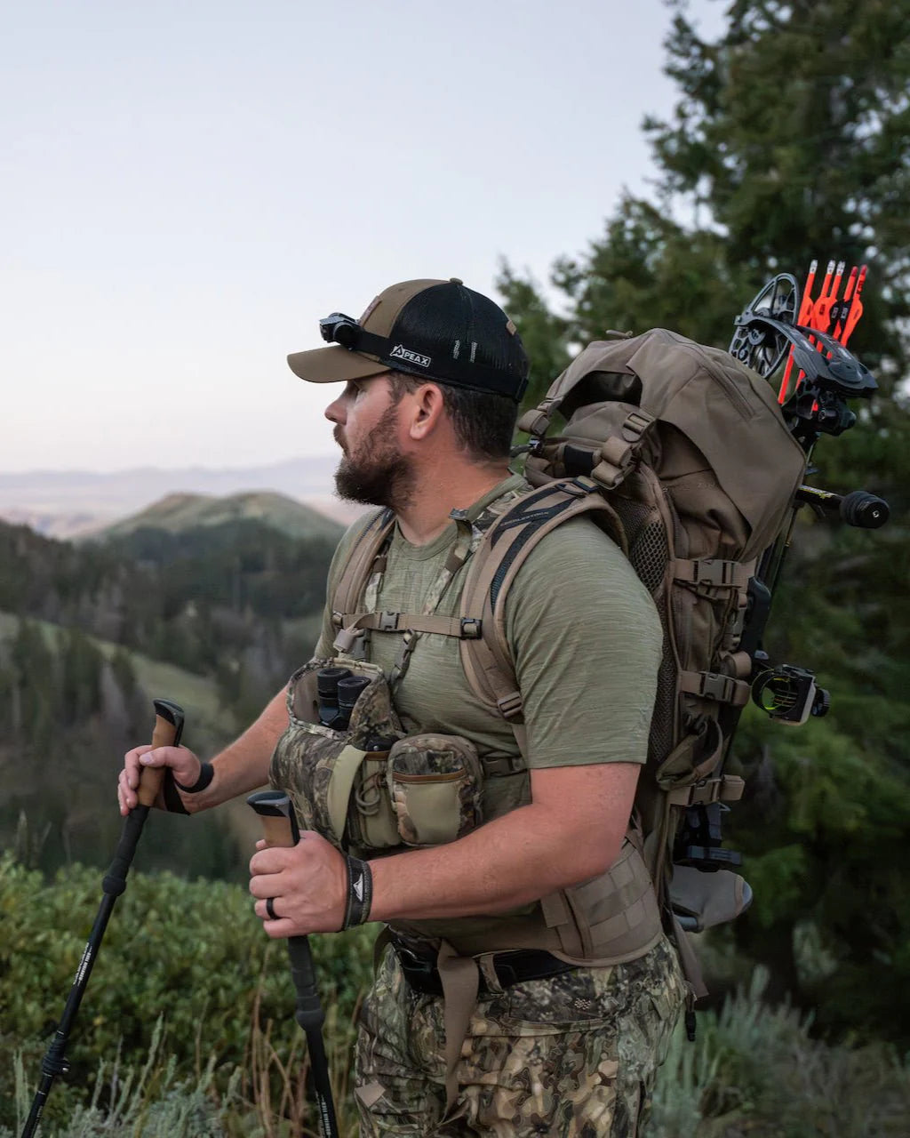 Ein Mann in militärgrüner Outdoor-Ausrüstung, der den ultraleichten EBERLESTOCK Vapor 2500 V2 Rucksack und eine Bogenschießausrüstung trägt, steht mit Trekkingstöcken auf einem baumbestandenen Hügel und blickt auf die fernen Berge.