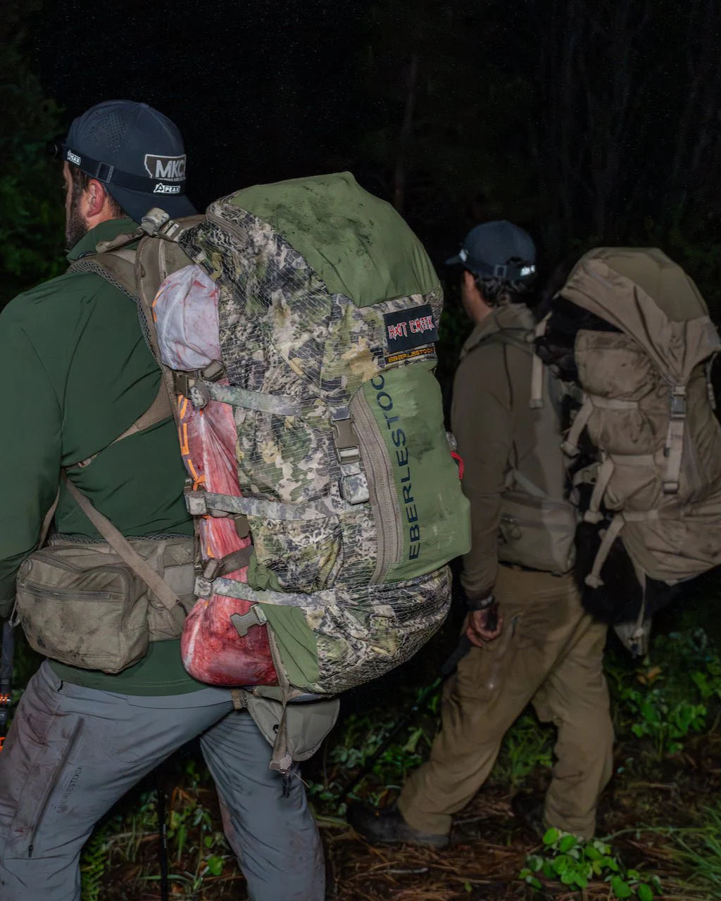 Zwei Wanderer mit großen EBERLESTOCK Rucksäcken, darunter der Eberlestock Vapor 2500 V2, laufen durch einen dunklen Wald. Auf einen Rucksack ist Wildfleisch geschnallt, während sie die Vegetation auf dem Waldboden passieren.