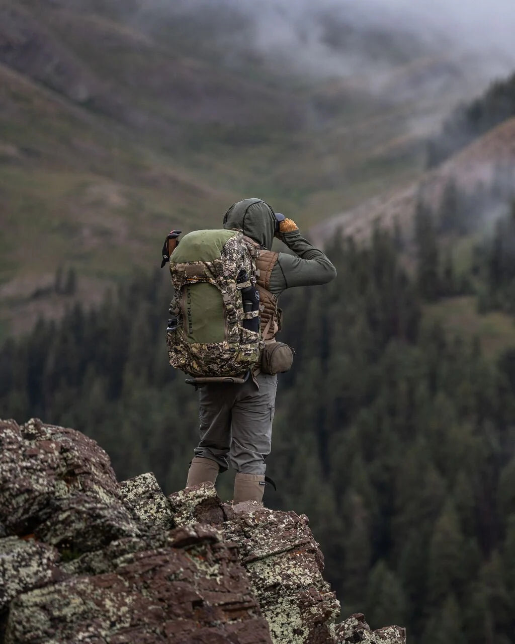 Bekleidet mit Outdoor-Ausrüstung und dem EBERLESTOCK Vapor 2500 V2 steht eine Person auf einem Felsvorsprung und blickt über eine neblige, bewaldete Berglandschaft.