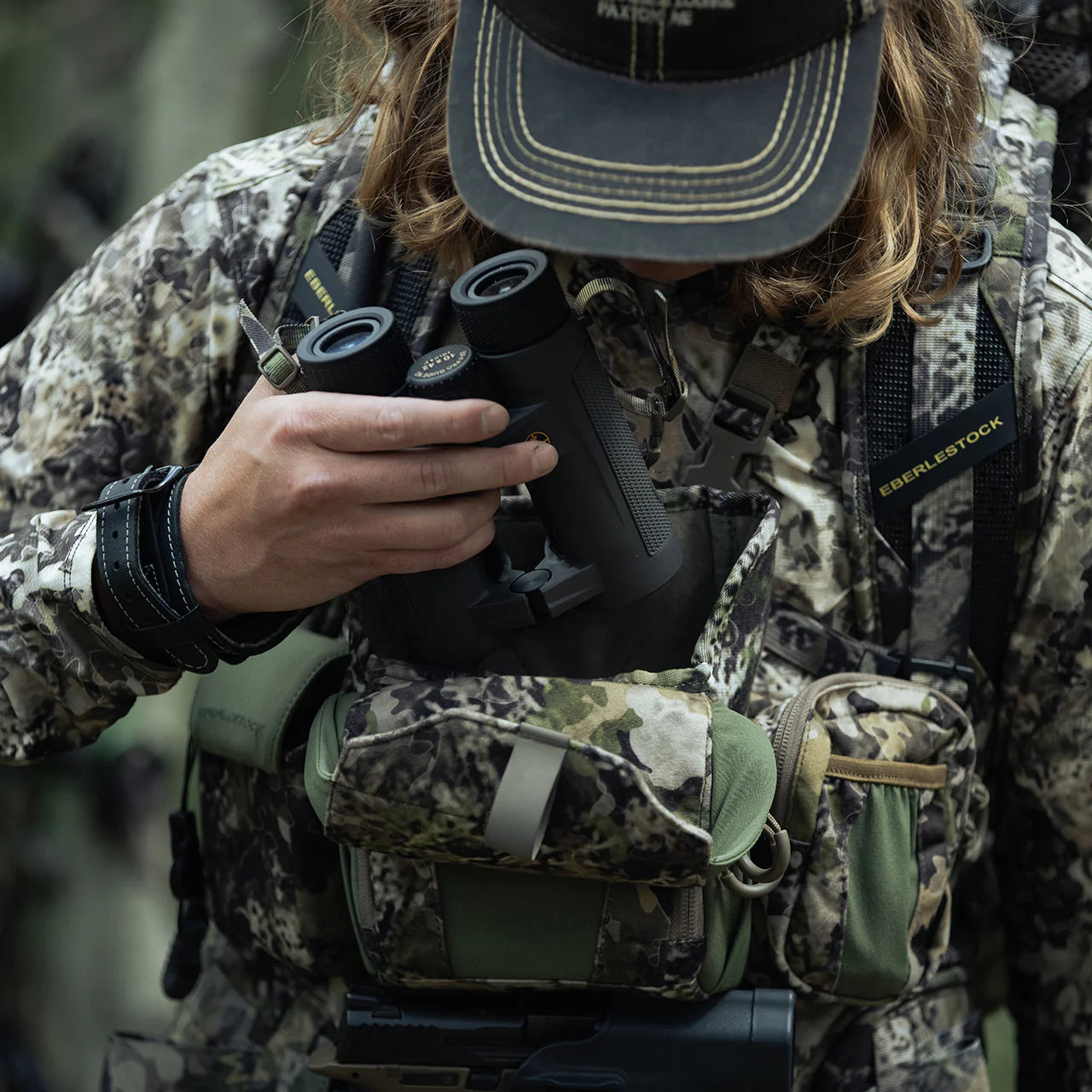Eine Person in Tarnkleidung und mit Hut hält im Freien ein Fernglas in der Hand, während sie sich das EBERLESTOCK Recon Modular Bino Pack Small und die Ausrüstung auf die Brust schnallt.