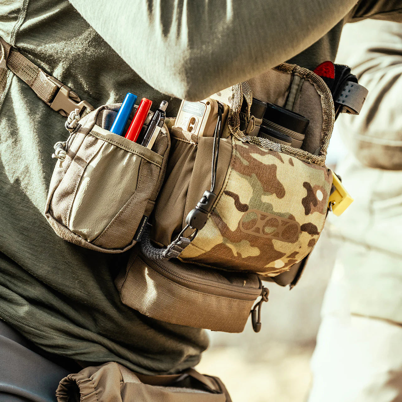 Ein taktischer Gürtel mit Tarnmuster und braunen Taschen für Stifte, Werkzeug und Ausrüstung passt gut zu einem grünen Outfit, ergänzt durch das EBERLESTOCK Recon Modular Bino Pack Small Fernglas-Tragesystem.