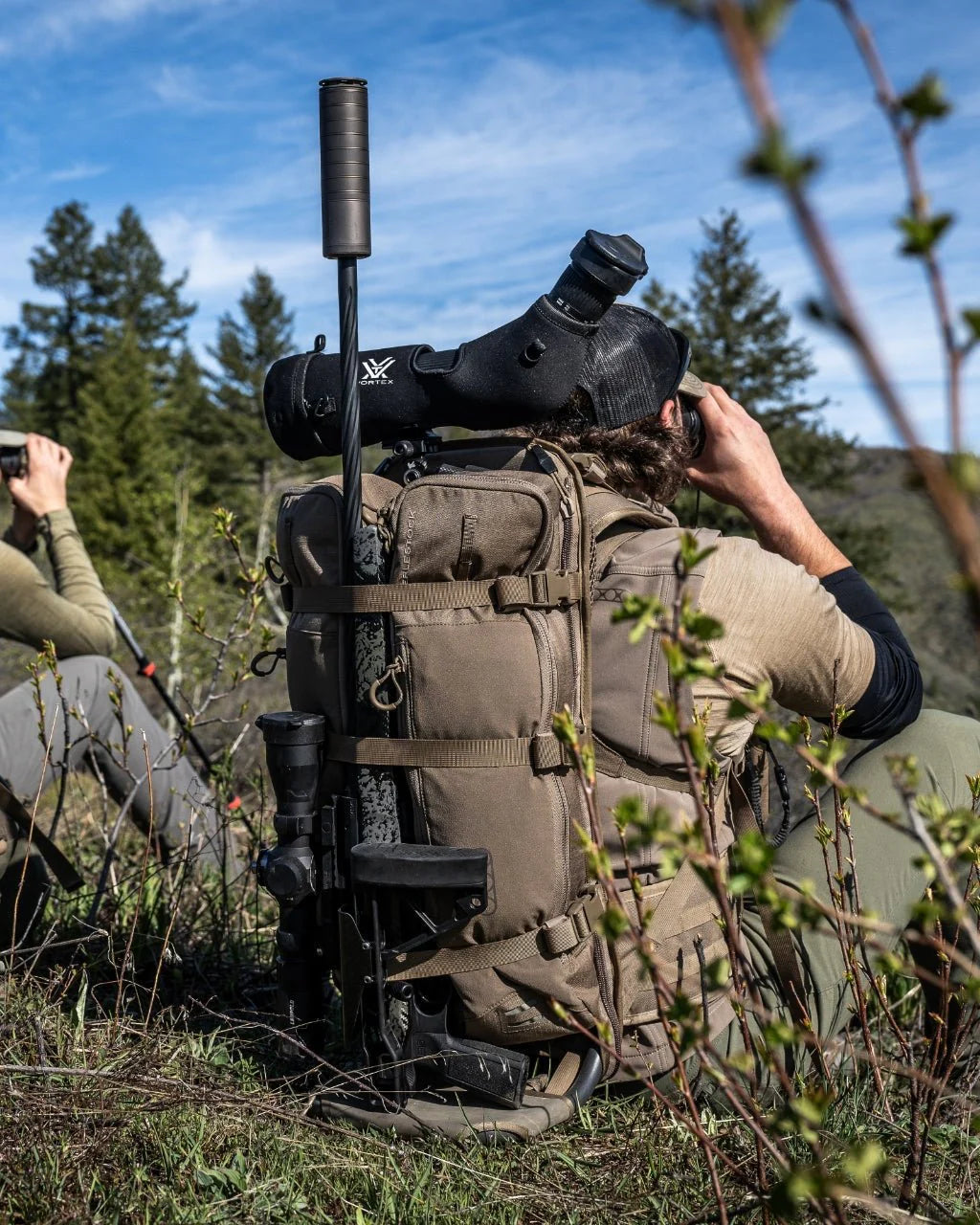 Zwei Personen in hellbrauner und grüner Kleidung sitzen im Freien auf einer Wiese. Eine von ihnen benutzt ein EBERLESTOCK Batwing V2 mit Stativ und Spektiv und schaut durch ein Fernglas inmitten von Bäumen unter einem blauen Himmel.