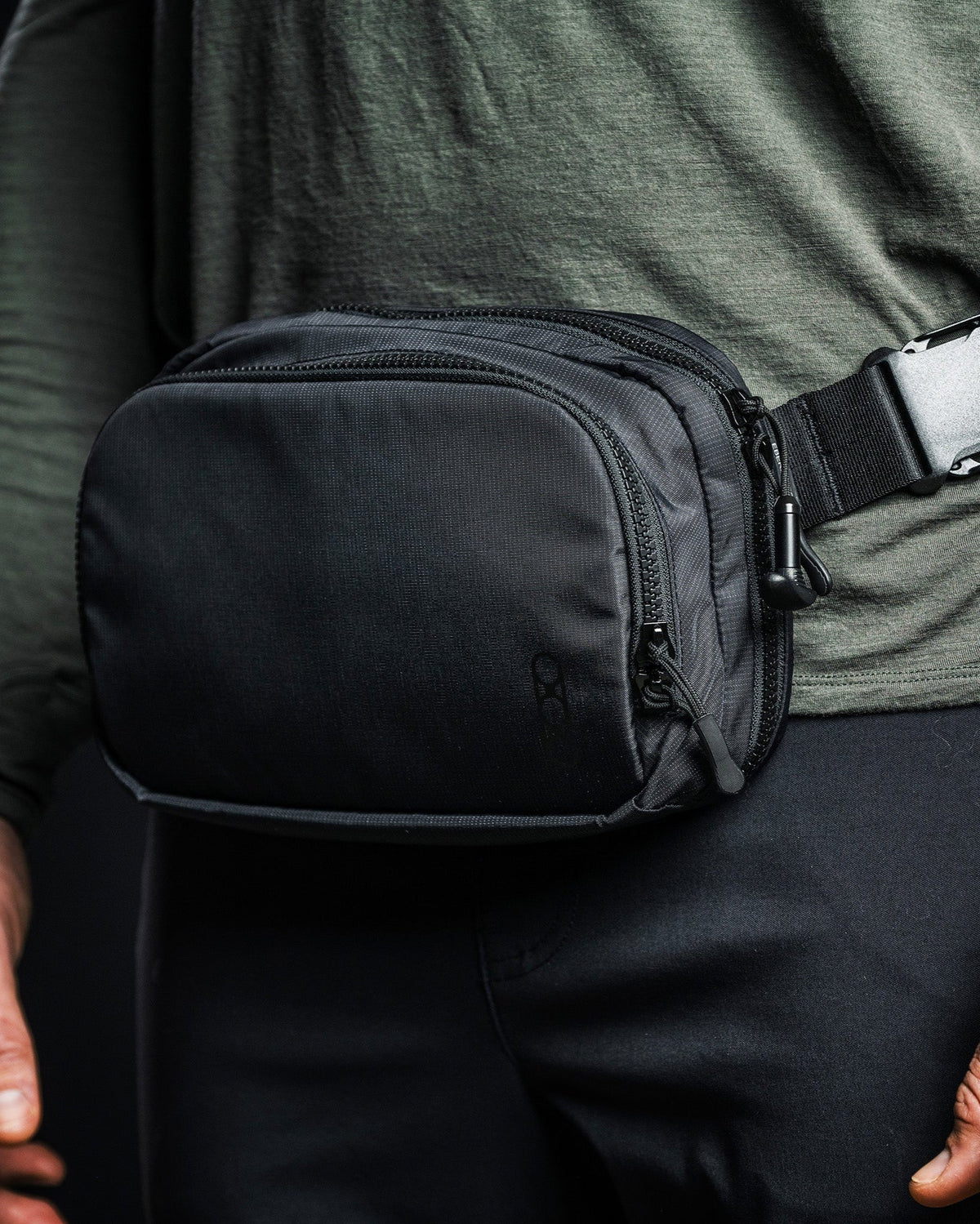 Eine Person in einem dunkelgrünen langärmeligen Hemd und einer dunklen Hose trägt die EBERLESTOCK Fade Flex Hüfttasche mit Schnallenverschluss und mehreren Fächern. Die Eberlestock Fade Flex kann auch als Crossbody-Tasche getragen werden.
