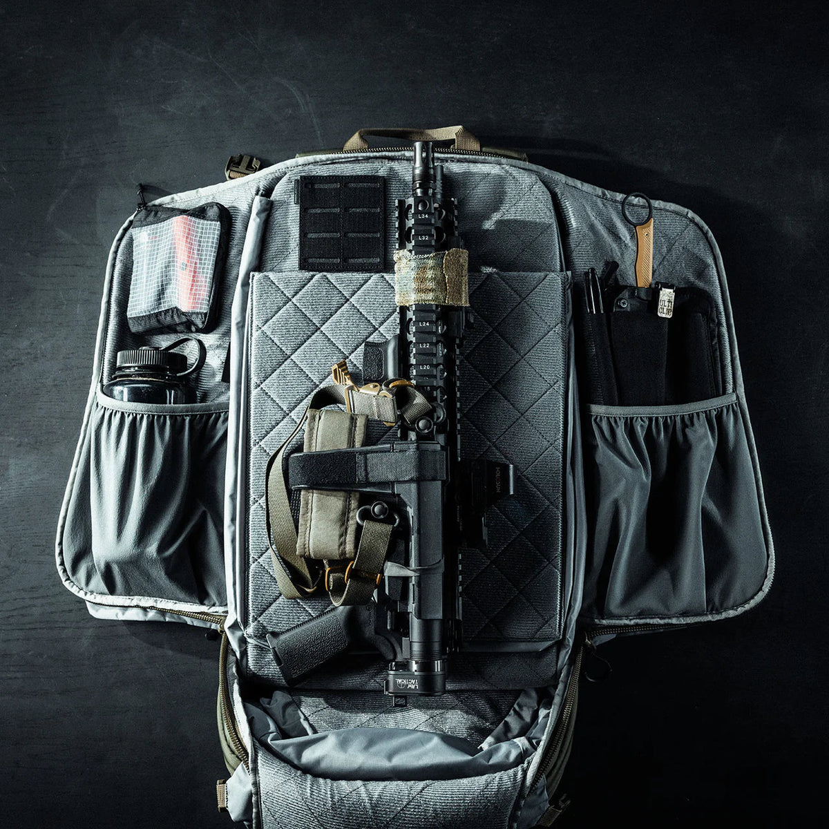 Ein taktischer EBERLESTOCK Rucksack Fade Adapt liegt geöffnet auf dunklem Untergrund. Im ausklappbaren Scabbard sind Gewehr, Pistole im Holster, Magazin, Tourniquet, Taschenlampe und weitere Ausrüstung ordentlich angeordnet.