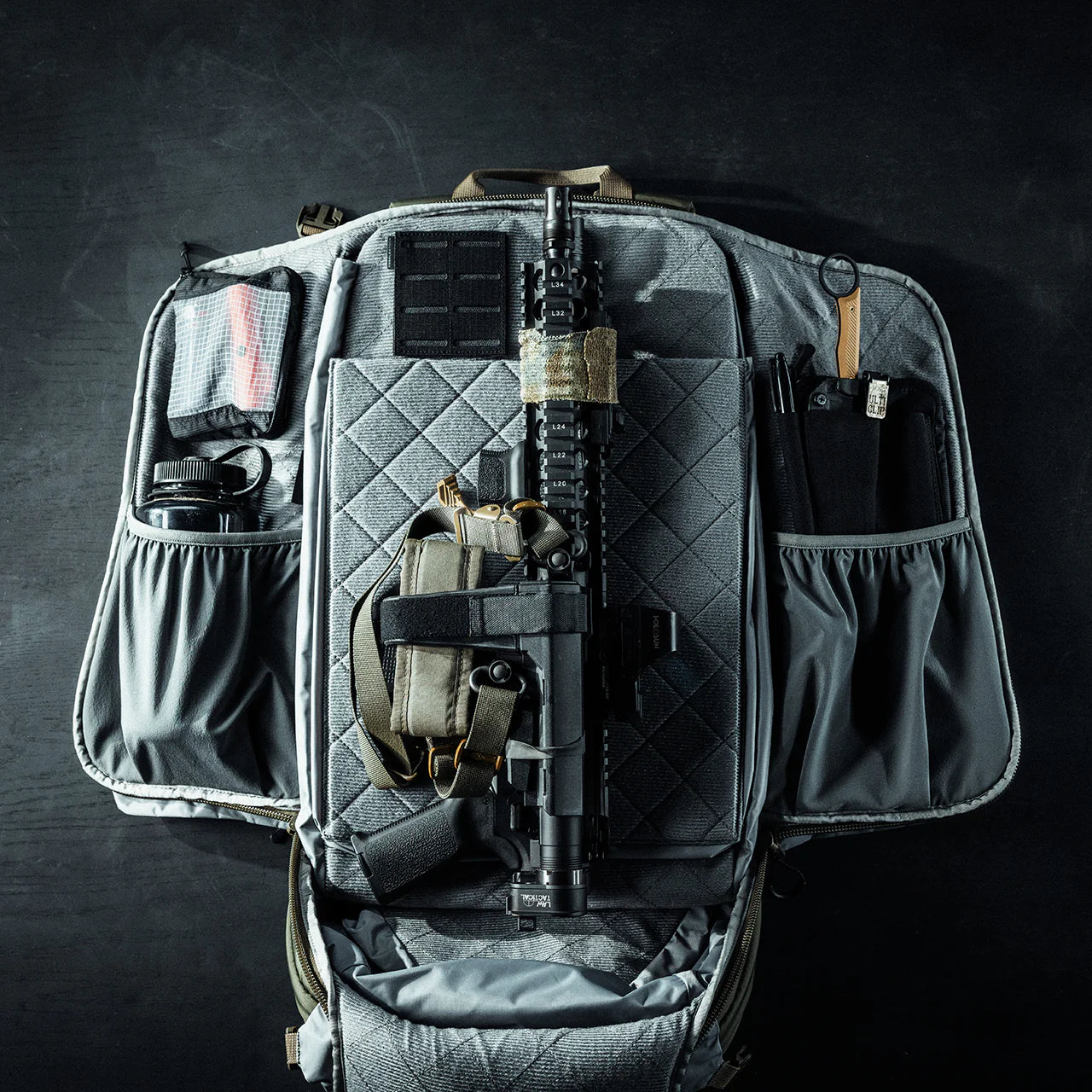 Ein taktischer EBERLESTOCK Rucksack Fade Adapt liegt geöffnet auf dunklem Untergrund. Im ausklappbaren Scabbard sind Gewehr, Pistole im Holster, Magazin, Tourniquet, Taschenlampe und weitere Ausrüstung ordentlich angeordnet.