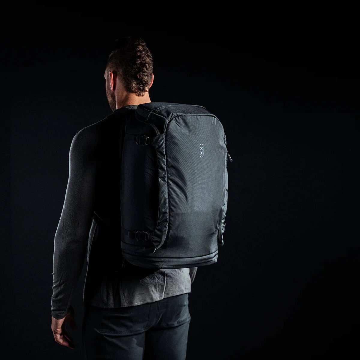 Eine Person, bekleidet mit einem dunklen, langärmeligen Hemd und mit dem Rücken zur Kamera, trägt den EBERLESTOCK Fade Adapt Rucksack vor einem schwarzen Hintergrund.