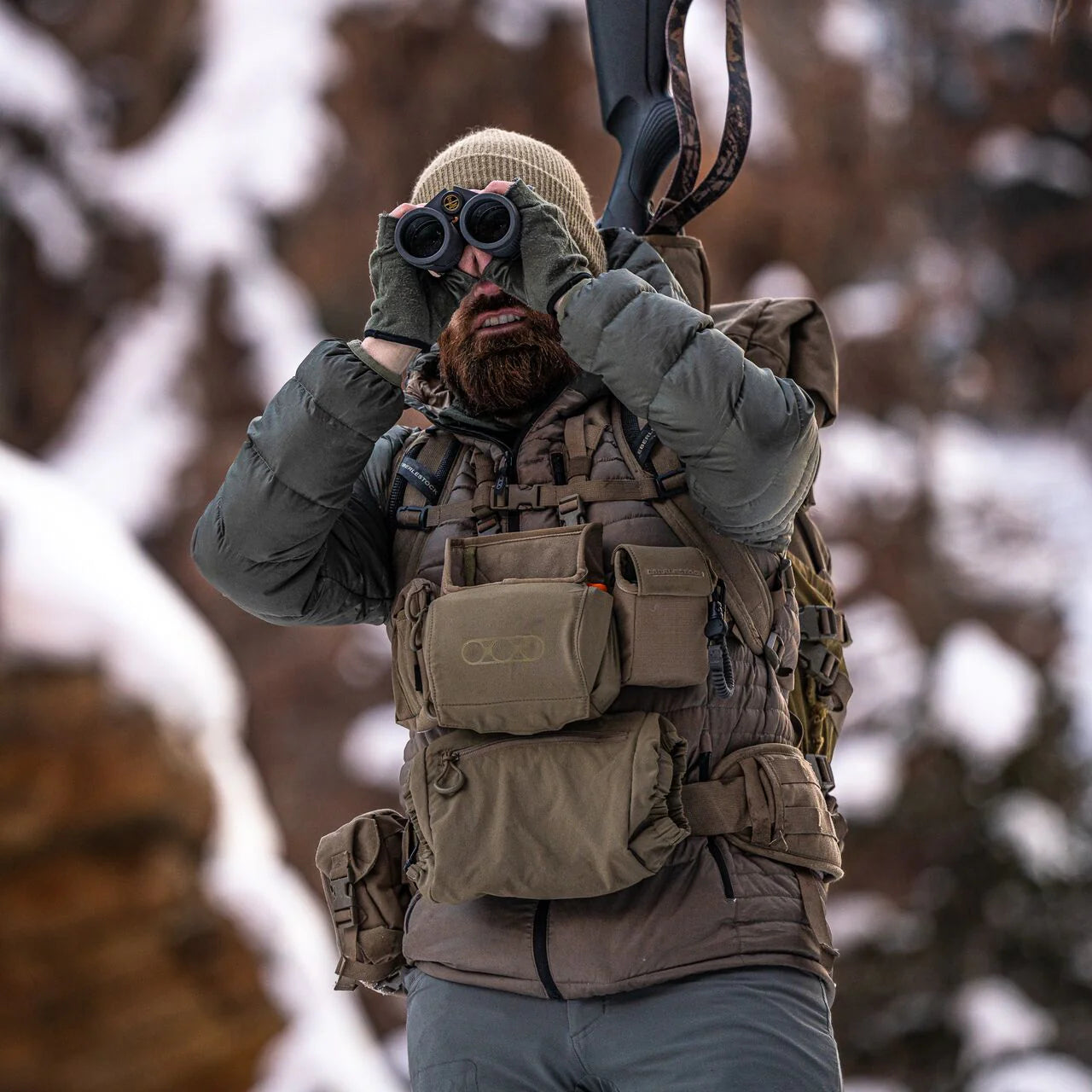 Ein bärtiger Mann in Winterkleidung und mit Mütze benutzt in verschneitem Gelände ein Fernglas. Er trägt ein Gewehr auf der Schulter und hat einen EBERLESTOCK Recon Modular Bino Pack Large Brustbeutel am Körper.