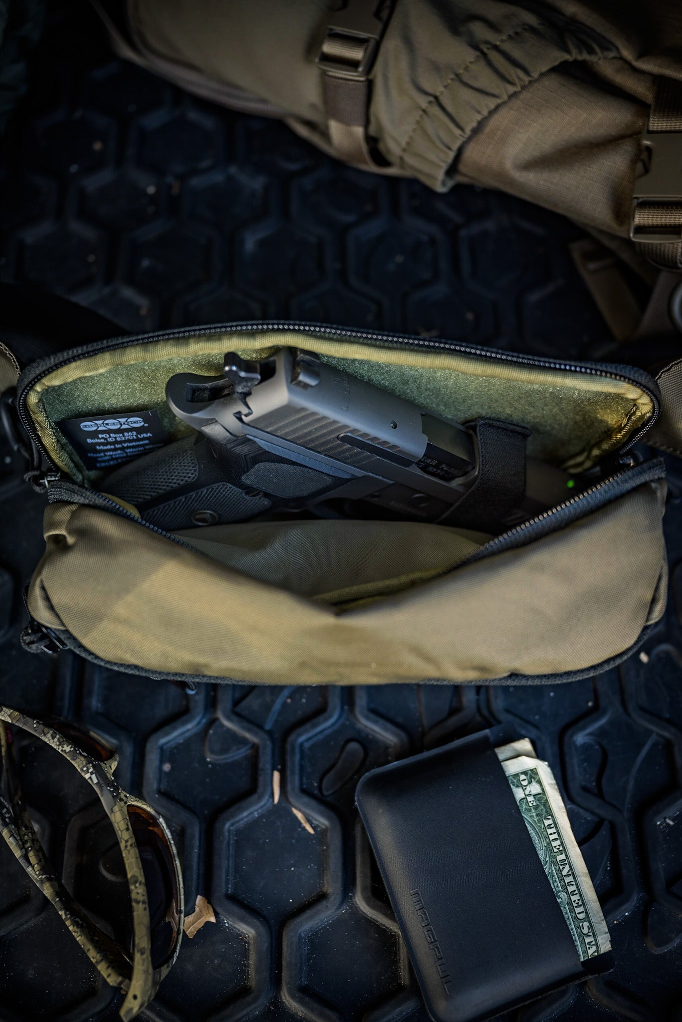 Eine geöffnete EBERLESTOCK Bando Bag Standard in Olivgrün zeigt eine Handfeuerwaffe in ihrem verdeckten CCW-Fach auf einer strukturierten schwarzen Oberfläche, daneben eine gefaltete Sonnenbrille und eine schwarze Brieftasche mit einem sichtbaren Dollarschein.