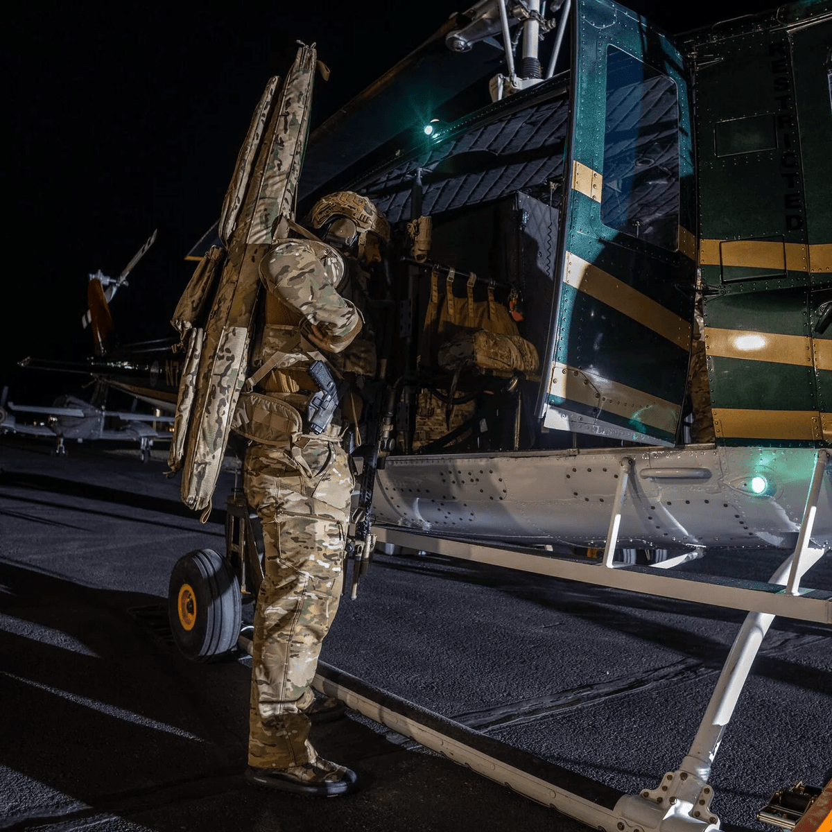 Ein Soldat in Tarnkleidung und mit Helm steht nachts neben einem Hubschrauber und hält eine EBERLESTOCK Sniper Sled Drag Bag 52" in der Hand, während die geöffnete Seitentür den Blick auf das schwach beleuchtete Rollfeld freigibt, und bereitet sich auf den Einstieg vor.