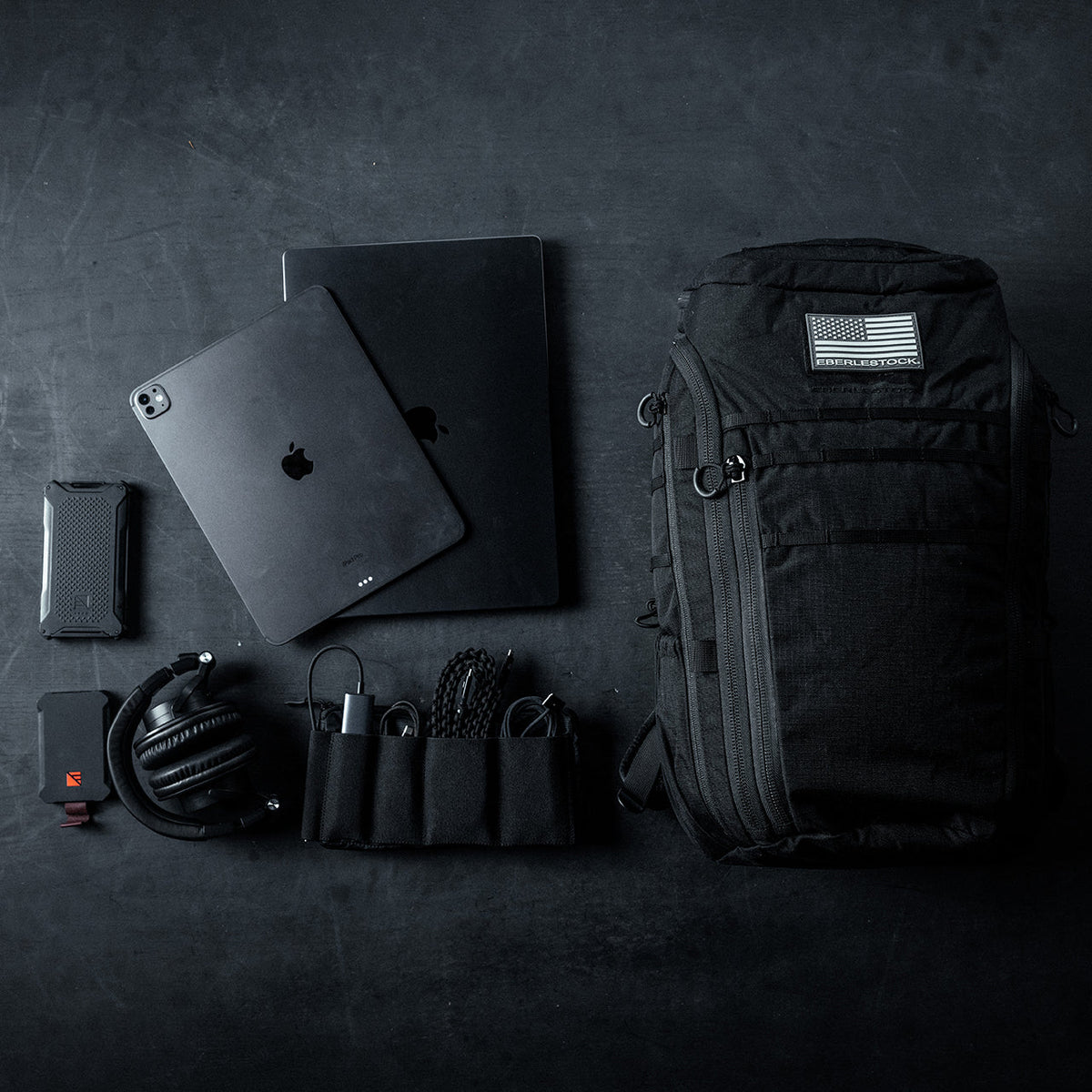 Eine flache Anordnung von schwarzem technischem Equipment und Zubehör, mit dem EBERLESTOCK Switchblade Pack mit einem Aufnäher der US-Flagge, einem iPad, MacBook, Kopfhörern, tragbaren Ladegeräten, Kabeln und einem kleinen Organizer auf einer dunklen Oberfläche.