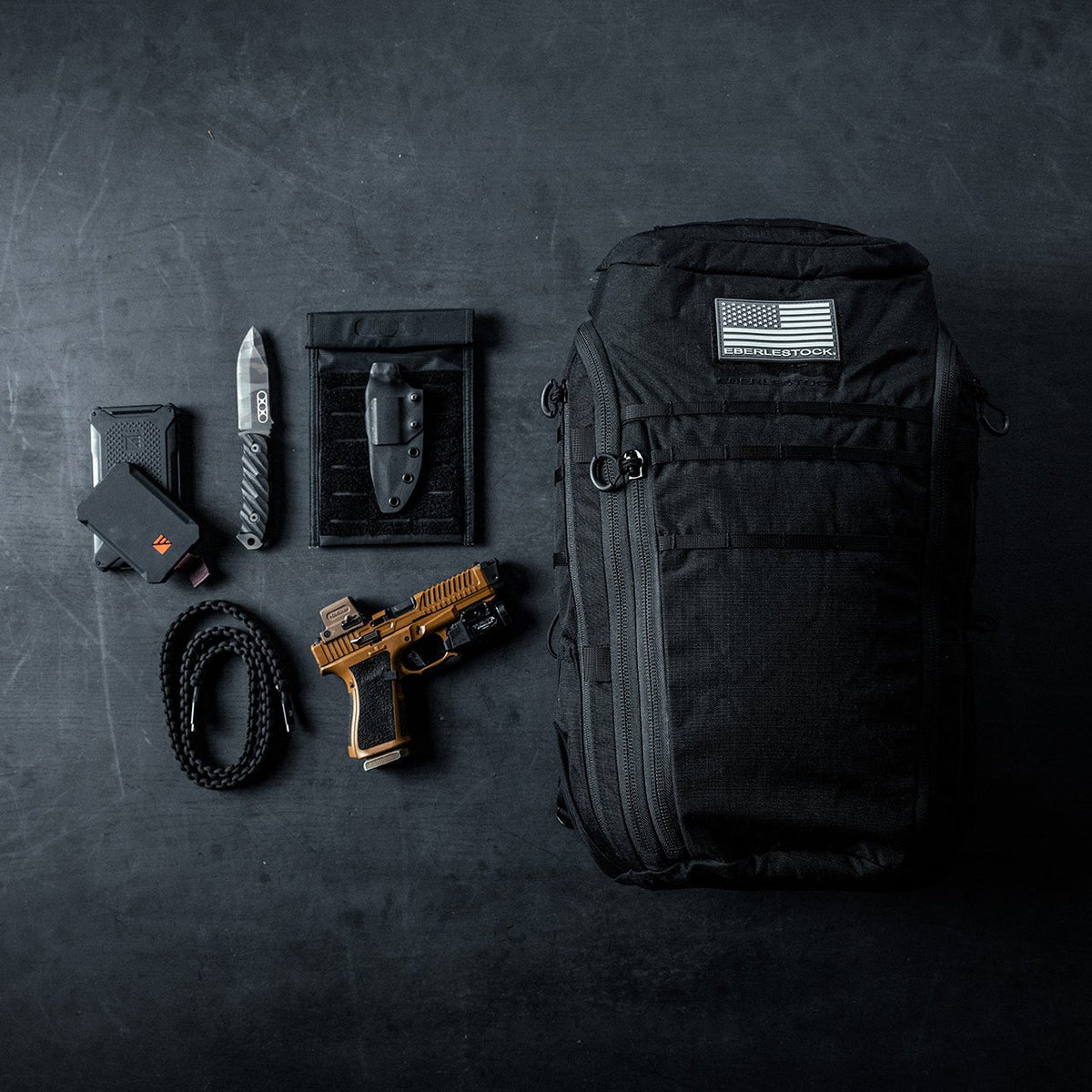 Ein schwarzer EBERLESTOCK Switchblade Pack mit einem Aufnäher der US-Flagge liegt auf einer dunklen Fläche, umgeben von einer Pistole, einem Klappmesser, einem Multitool, einem aufgerollten Seil und einem kleinen schwarzen Etui - ideal für alle, die eine elegante taktische Rucksackausrüstung suchen.