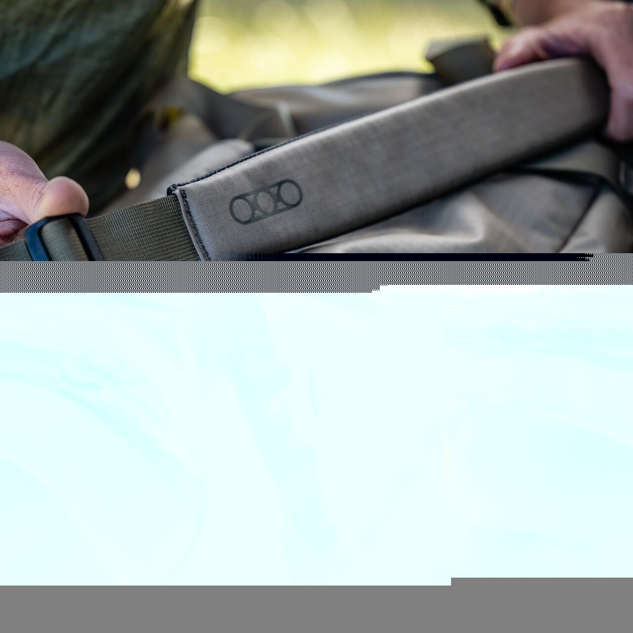 Eine Person hält den Gurt einer EBERLESTOCK Hyllus Duffel 65, einer 65-Liter-Tasche, im Freien bei natürlichem Licht. Der untere Teil des Bildes ist digital verzerrt.