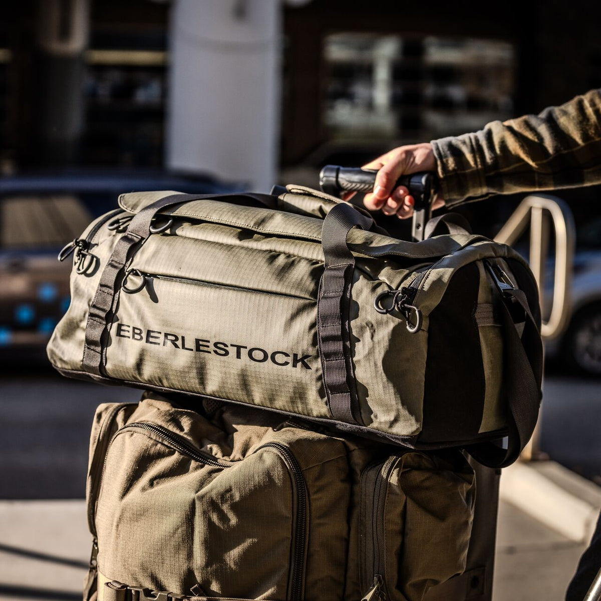 Eine Person hält den Griff eines EBERLESTOCK Hyllus Duffel 65 in khakigrün, der auf einer passenden Tasche im Freien im Sonnenlicht vor einer unscharfen Stadtkulisse steht.