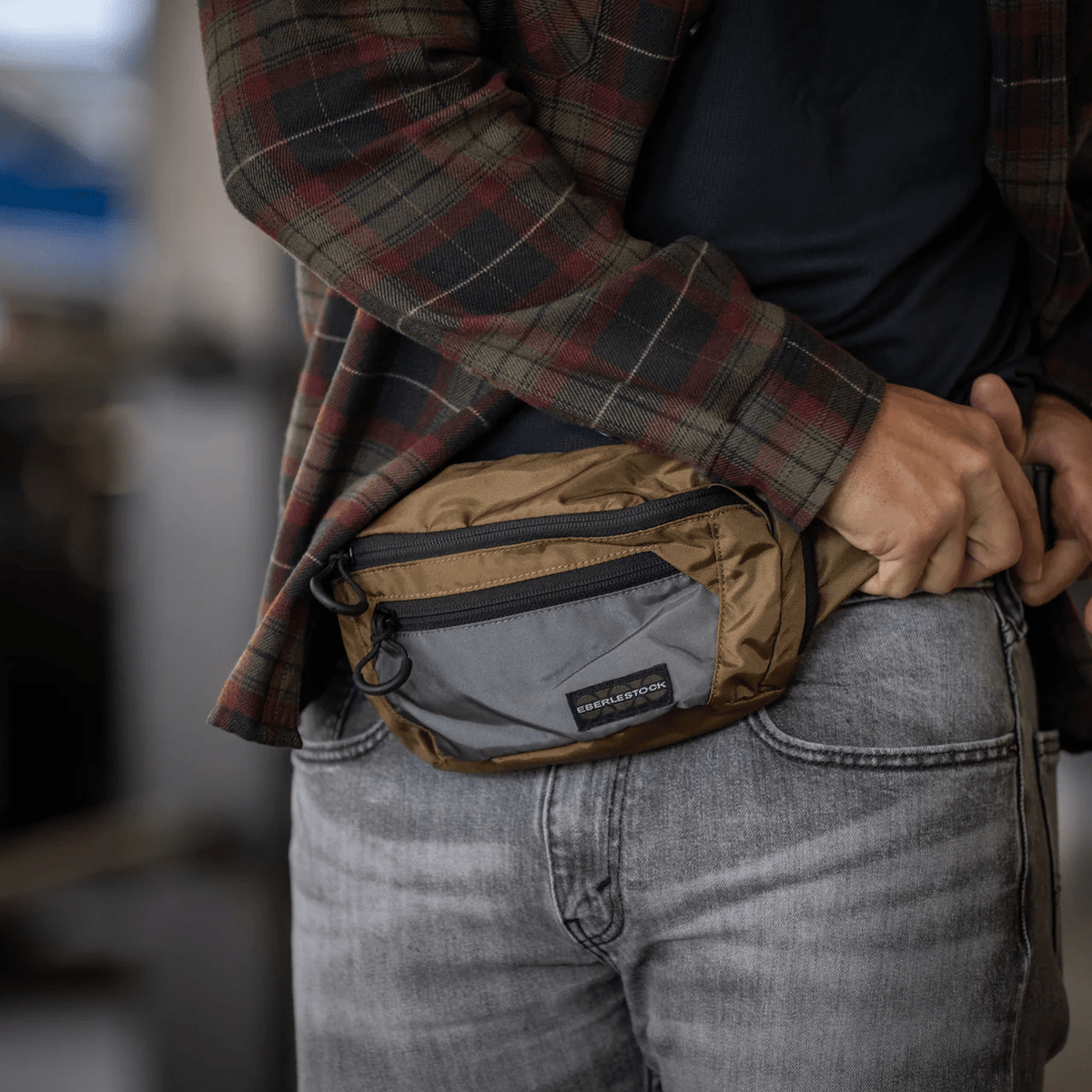 Eine Person, die ein kariertes Hemd und eine Jeans trägt, passt die EBERLESTOCK Bando Bag Standard - perfekt für das verdeckte Tragen - mit einer Reißverschlusstasche und einem kleinen schwarzen Etikett an, während sie in einem Haus in einer legeren Umgebung steht.