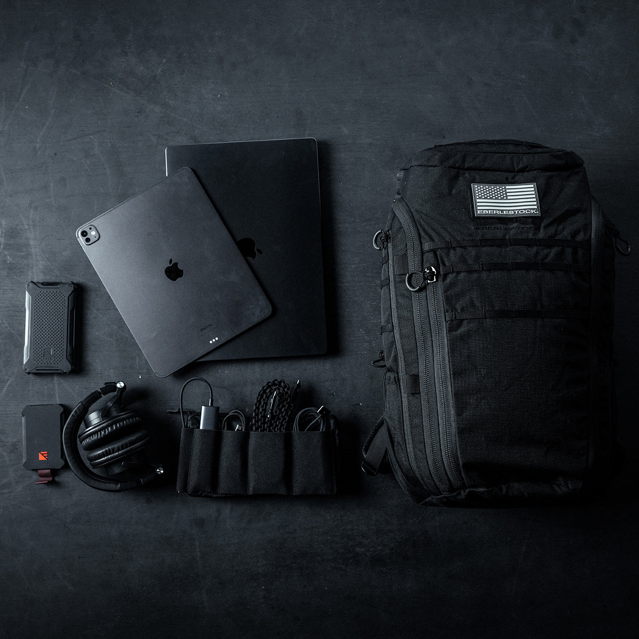 Flachdruck von schwarzer Technik- und Reiseausrüstung auf einer dunklen Oberfläche, mit dem EBERLESTOCK Switchblade Pack aus robustem 500D-Nylon, abgebildet mit Laptop, Tablet, Smartphone, Kopfhörer, tragbarem Ladegerät, Kabeln, Netzadapter und Zubehörtasche.