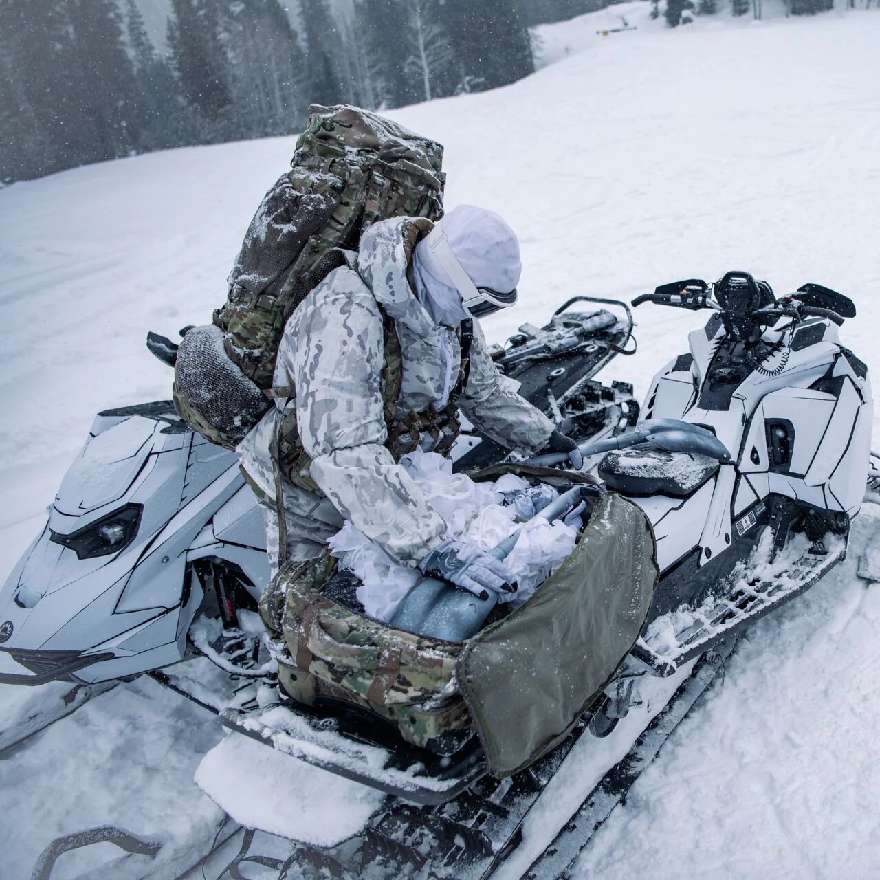 Eine Person in weißer und getarnter Winterkleidung öffnet einen EBERLESTOCK Atlas Duffel auf dem Sitz eines Schneemobils in einem verschneiten Wald, während ein anderes Schneemobil in der Nähe ist.