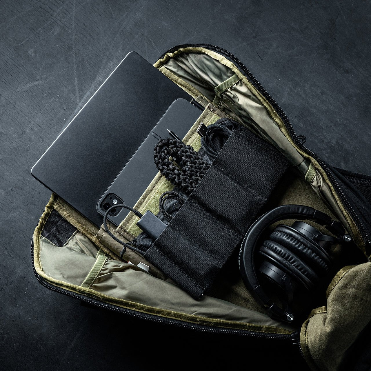 Ein geöffneter EBERLESTOCK Switchblade Pack aus strapazierfähigem 500D-Nylon liegt auf einer dunklen Oberfläche und zeigt Laptop, Smartphone, Kopfhörer, Ladegerät und organisierte Kabel in seinen gepolsterten taktischen Fächern.