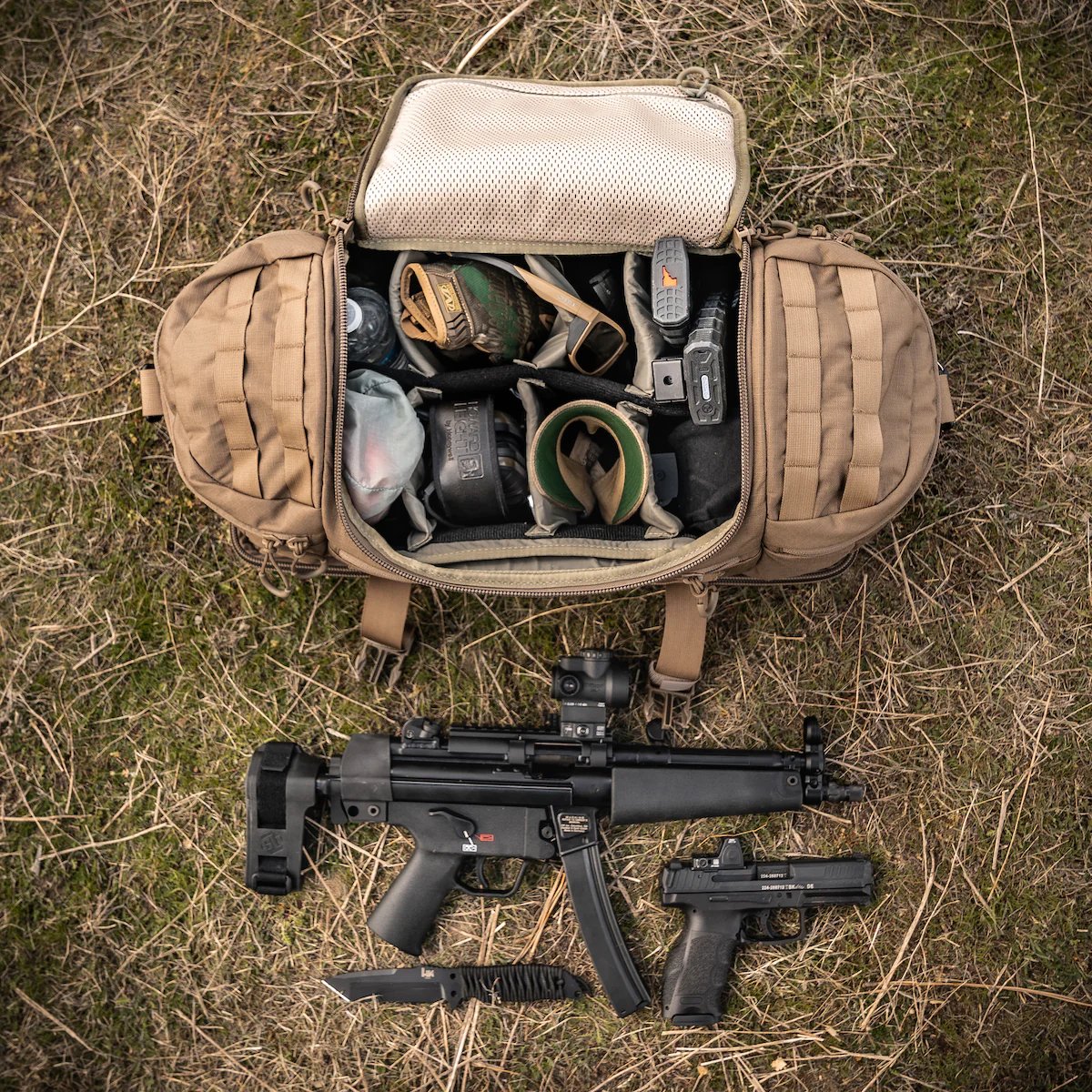 Ein EBERLESTOCK Bang Bag in hellbraunem, robustem 500D-Nylon liegt auf dem Gras, mit offenen Fächern für Handschuhe, Schutzbrille und Ausrüstung. Auf der Vorderseite liegen eine schwarze Maschinenpistole, eine Pistole, ein Kampfmesser und ein Magazin.