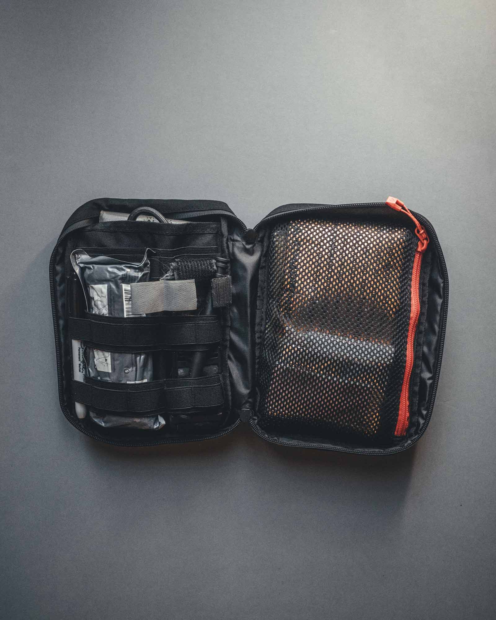Ein offenes schwarzes STOIRM Large Pouch V.2 Erste-Hilfe-Kit mit organisiertem Zubehör auf der linken Seite und weiteren Gegenständen hinter einer Netztasche mit rotem Reißverschluss auf der rechten Seite, vor einem grauen Hintergrund - kompatibel mit Molle-Panel-Systemen.