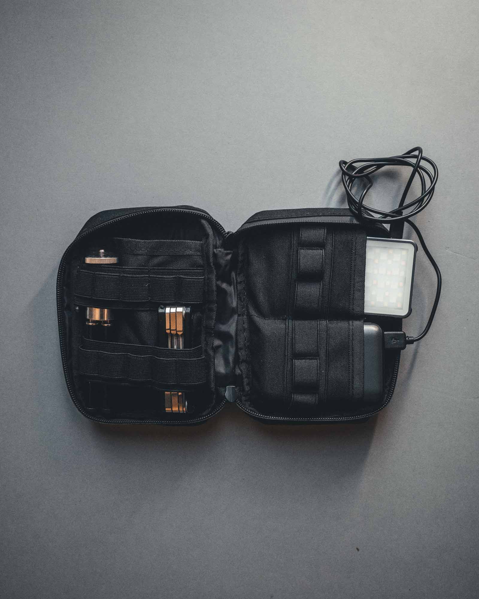 Eine geöffnete STOIRM Large Pouch V.1 von STOIRM zeigt ordentlich angeordnetes technisches Zubehör - Kabel, ein kleines LED-Panel und Gadgets - auf einem grauen Hintergrund, was sie zur perfekten vielseitigen kleinen Tasche für Ihre elektronischen Essentials macht.