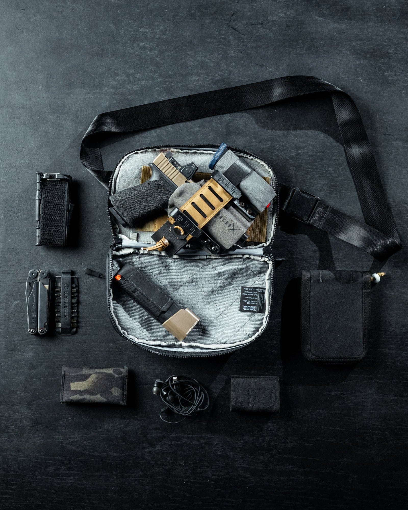 Eine EBERLESTOCK Eberlestock Fade Flex Crossbody-Tasche liegt offen auf einer dunklen Fläche und zeigt eine goldene und schwarze Handfeuerwaffe, ein Multitool und eine Ausrüstung. Um sie herum sind eine Taschenlampe, Kopfhörer, eine Brieftasche und Taschen angeordnet.