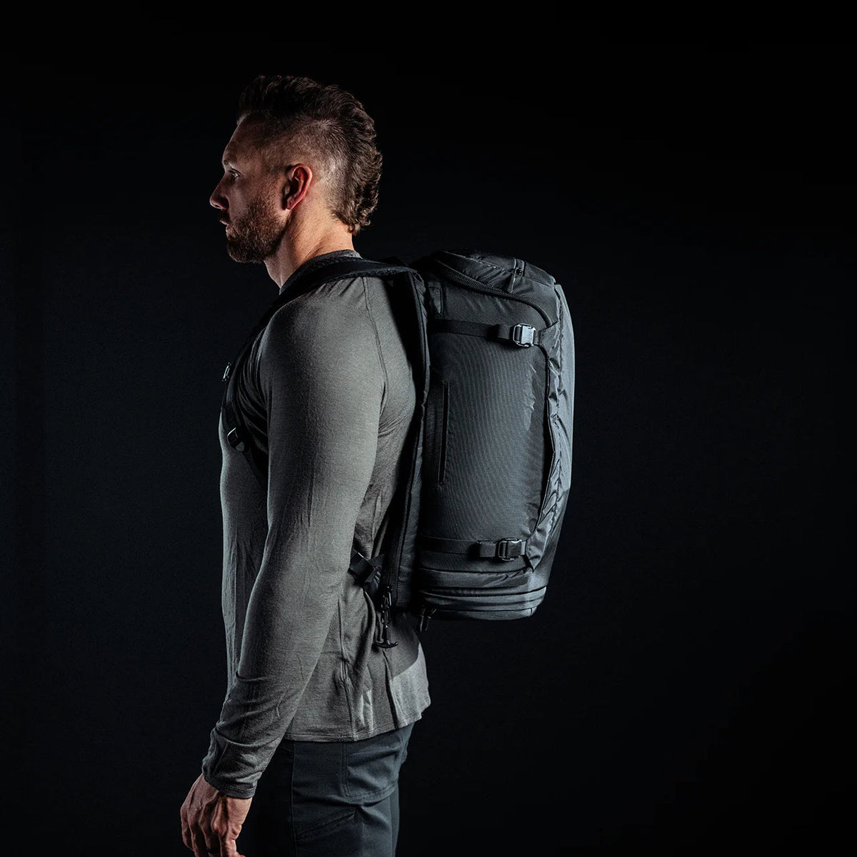 Ein Mann mit kurzem Bart, grauem langärmeligem Hemd und schwarzer Hose steht im Profil vor einem schwarzen Hintergrund und trägt den EBERLESTOCK Fade Adapt Rucksack mit aufklappbarer Scheide.