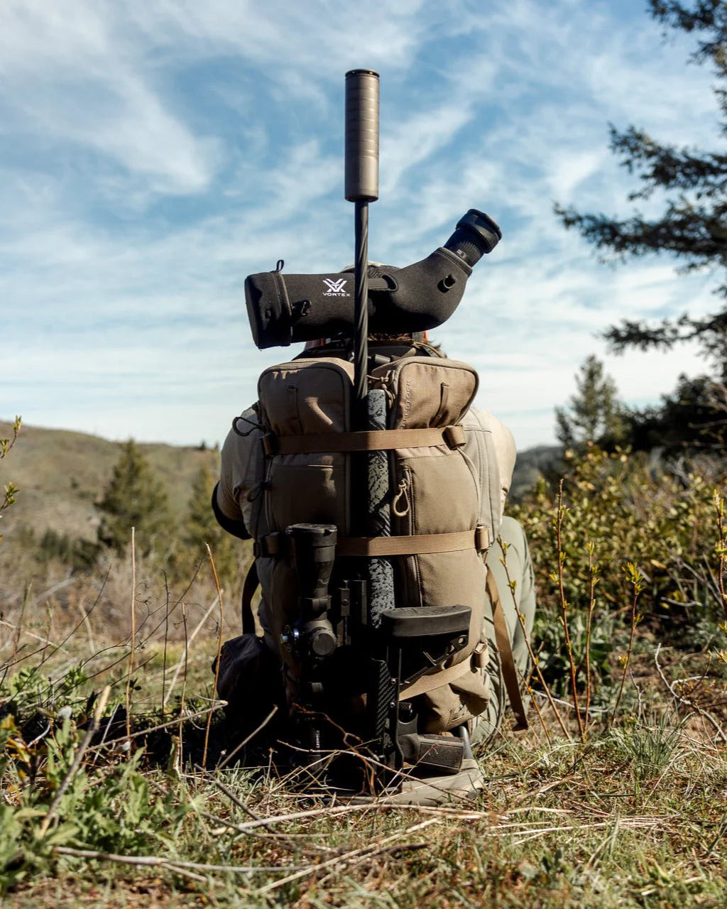Eine Person mit einem braunen Rucksack, der mit dem EBERLESTOCK Batwing V2 aus dem EMOD-System ausgestattet ist, sitzt mit einem Gewehr und einem Spektiv im Gras und blickt in die Ferne, umgeben von Bäumen und Hügeln unter einem blauen, wolkenverhangenen Himmel.