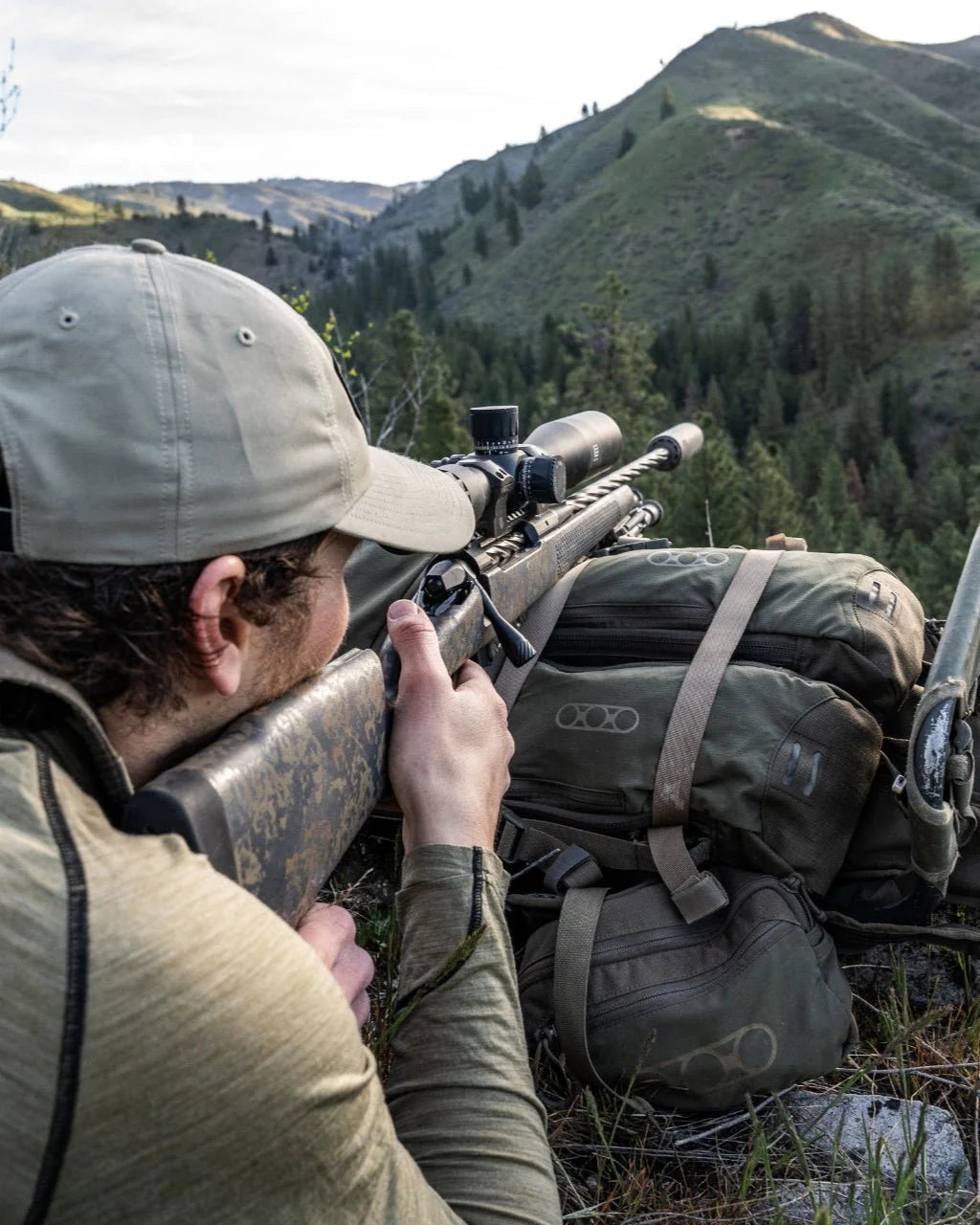 Eine Person in Outdoor-Ausrüstung liegt in Bauchlage auf einem Hügel und zielt mit einem Zielfernrohr auf ein Gewehr, daneben steht ein EBERLESTOCK Eberlestock Batwing V2, mit Blick auf eine bewaldete Berglandschaft.
