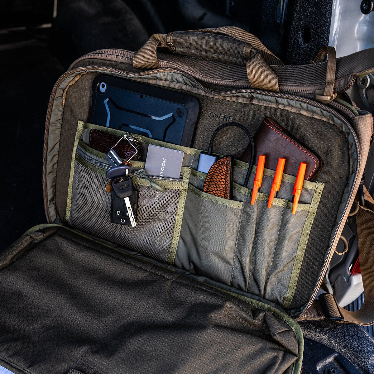 Eine geöffnete EBERLESTOCK Executive Brief zeigt organisierte Fächer für ein iPad, Schlüssel, ein Klappmesser, orangefarbene Stifte, ein Lederportemonnaie und eine braune gewebte Tasche auf einem Autositz - geräumig genug für einen 17-Zoll-Laptop.