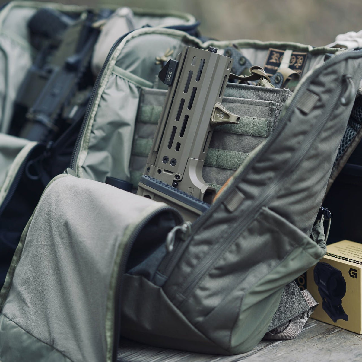 Das aus strapazierfähigem 500D-Nylon gefertigte EBERLESTOCK Switchblade Pack ist geöffnet und zeigt ein kompaktes Gewehr mit einem braunen Handschutz und Zubehör. Eine weitere Schusswaffe und Outdoor-Ausrüstung sind im Hintergrund teilweise sichtbar.