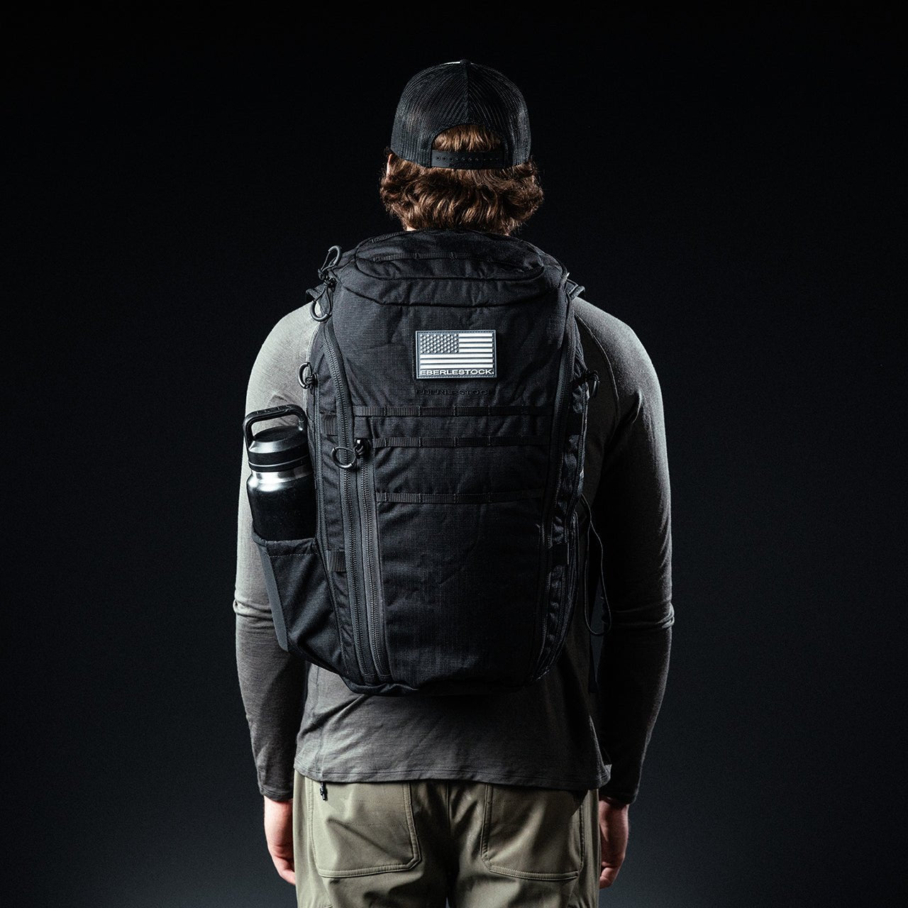 Eine Person mit schwarzer Mütze und grauem langärmeligem Hemd steht mit einem EBERLESTOCK Switchblade Pack - einem großen taktischen Rucksack aus 500D-Nylon mit einem Aufnäher der amerikanischen Flagge - und einer Wasserflasche vor einem dunklen Hintergrund.