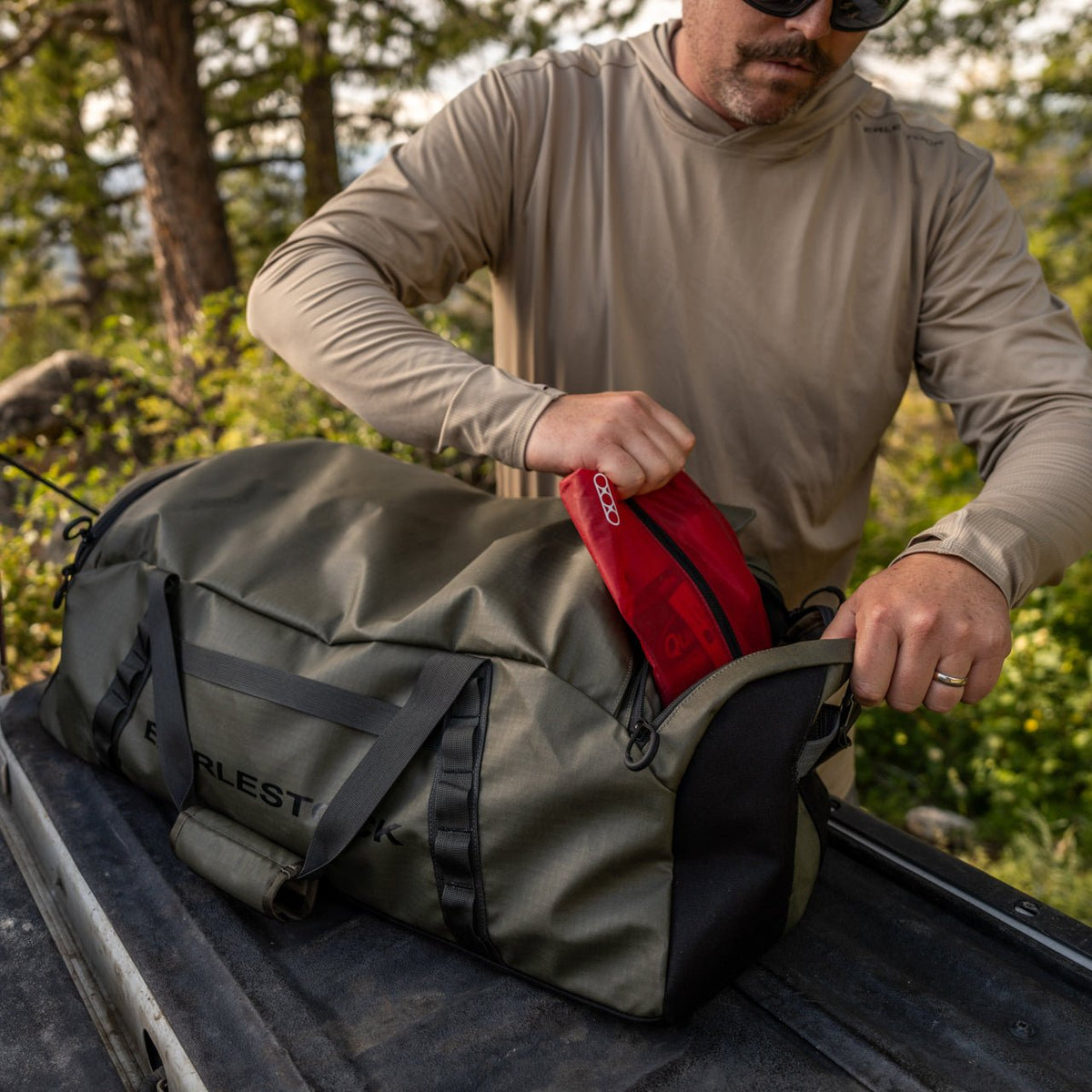 Ein Mann mit Sonnenbrille und beigefarbenem langärmeligem Hemd packt einen roten Gegenstand in einen EBERLESTOCK Hyllus Duffel 65 auf der Heckklappe eines Lastwagens, draußen im Sonnenlicht mit Bäumen im Hintergrund.