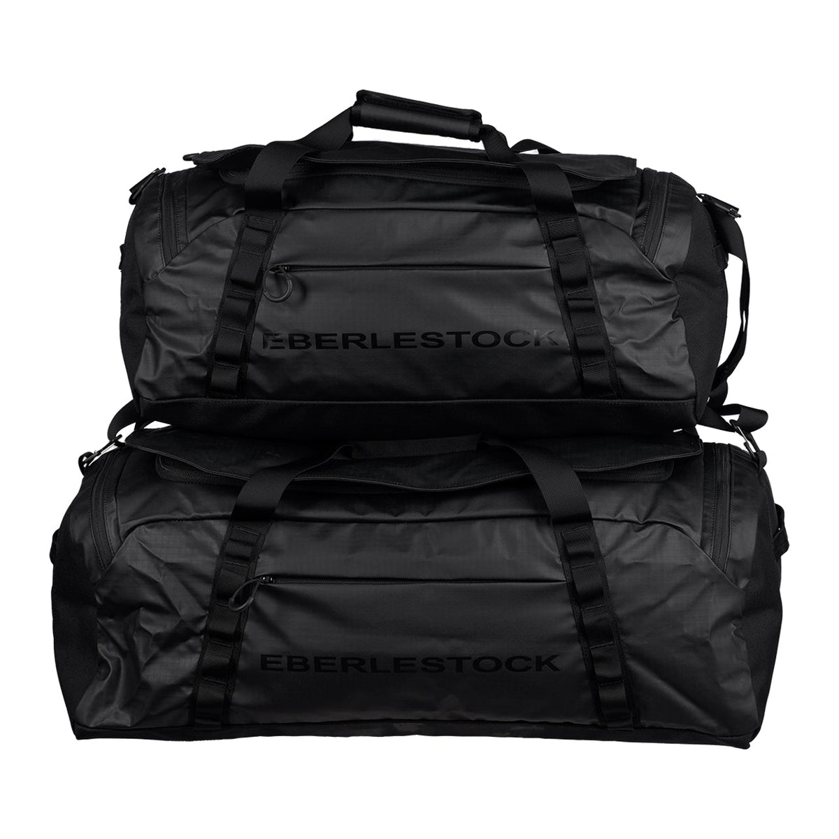 Zwei schwarze Eberlestock Hyllus Duffel 65 Taschen mit einem Fassungsvermögen von je 65 Litern sind mit stabilen Griffen und sichtbaren Reißverschlüssen gestapelt. Der markante EBERLESTOCK Markenname ist auf der Seite dieser vielseitigen Seesäcke aufgedruckt.